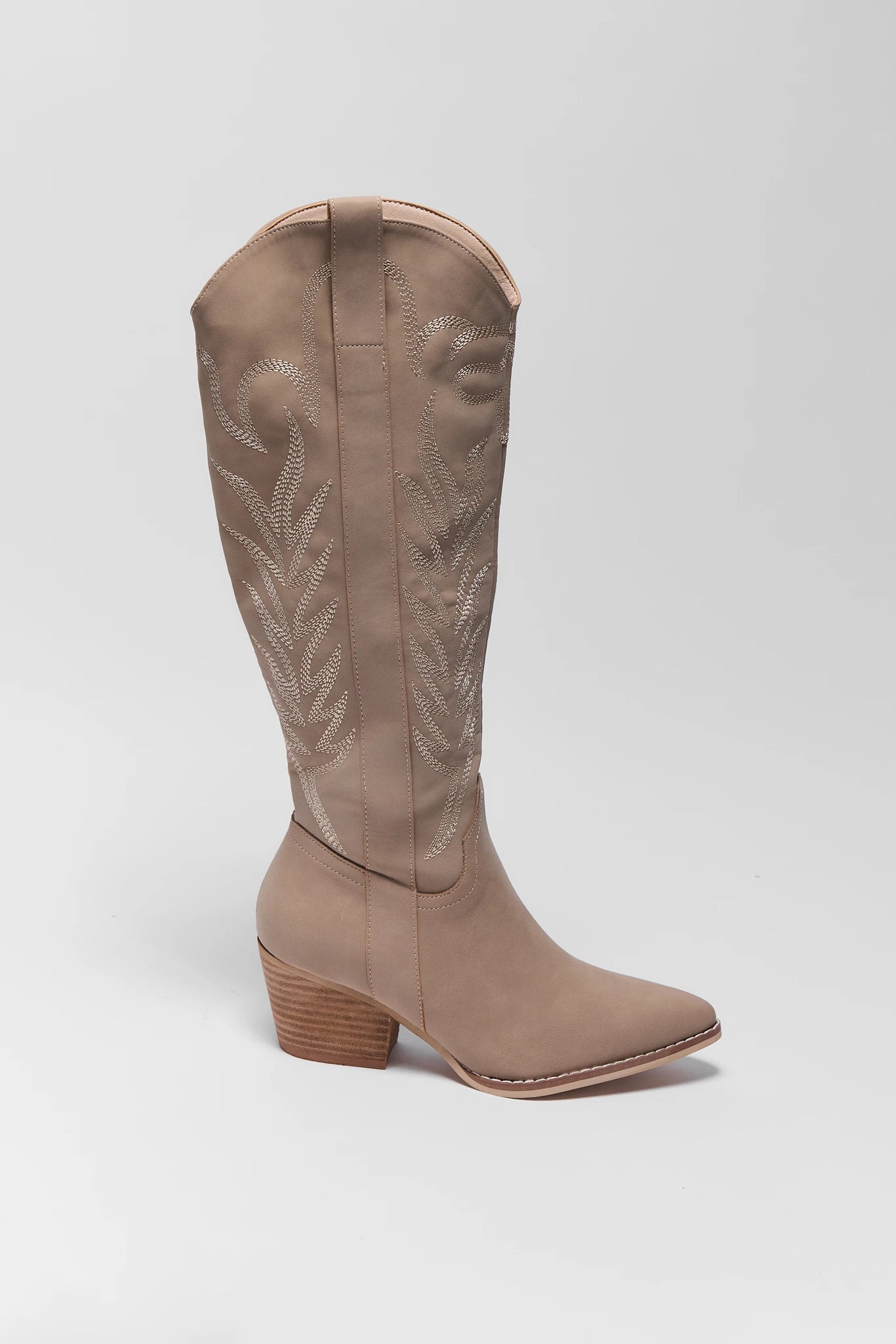 A single knee-high western boot in taupe suede with decorative cutout embroidery on the shaft, a pointed toe, and a stacked wood-look heel, shown against a light gray studio background.