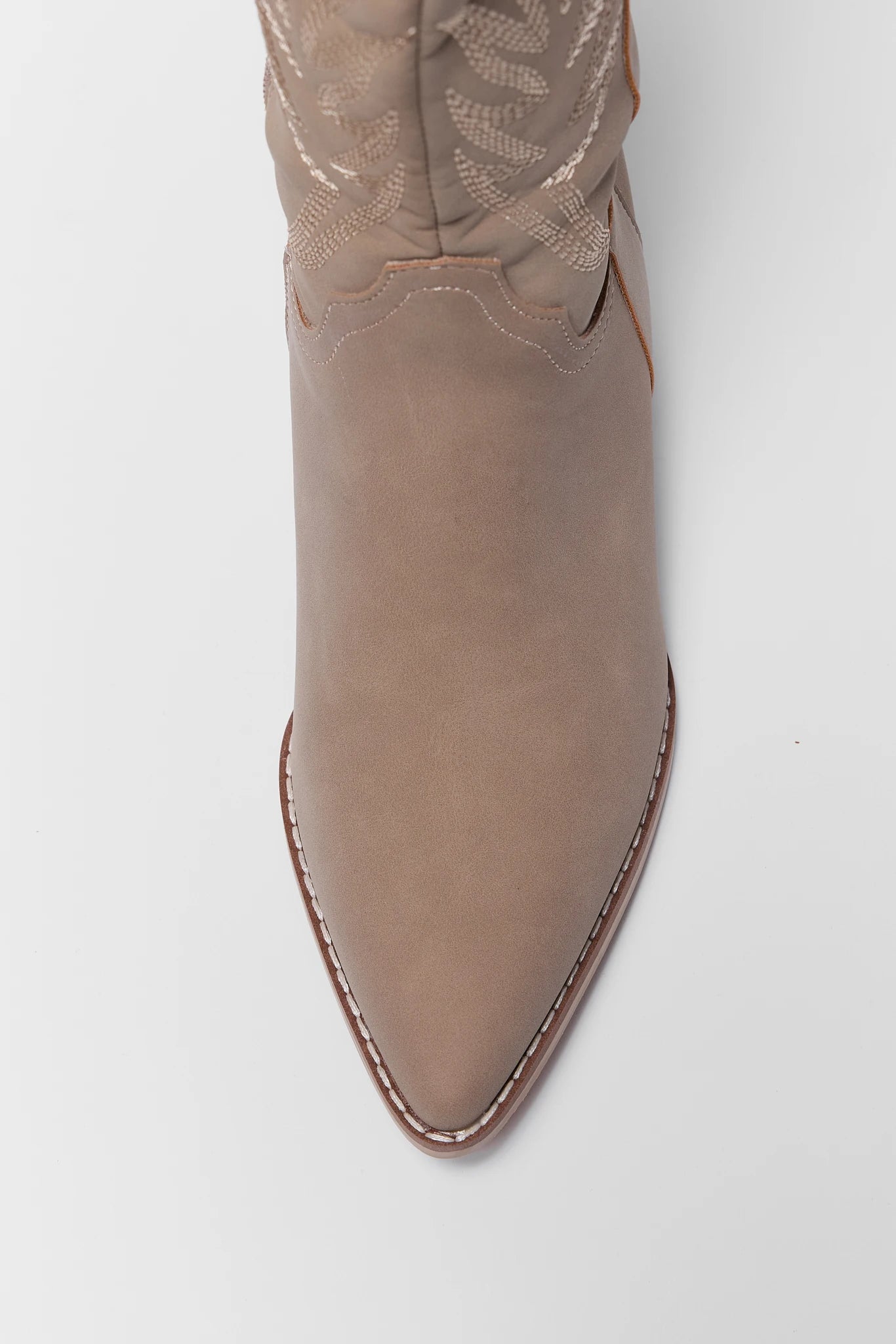 A close-up top-down view of a pointed-toe western boot in a taupe-beige suede-like finish, featuring decorative contrast stitching and a textured embroidered shaft on a light gray background.