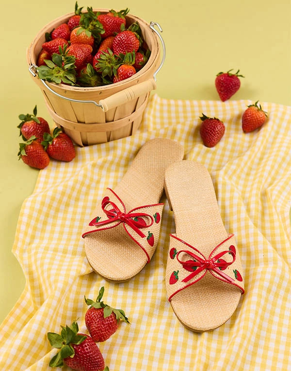 Beige sandals with red strawberry details on a yellow checkered tablecloth with strawberries around.