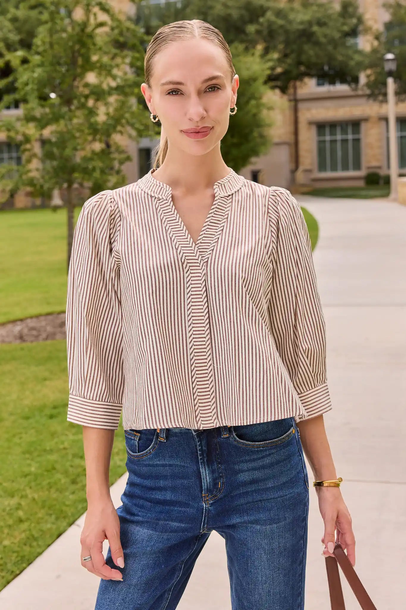 A woman wearing a beige and white vertically striped button-up blouse with puffed three-quarter sleeves, paired with blue jeans, standing outdoors on a paved walkway with trees and a building in the background.