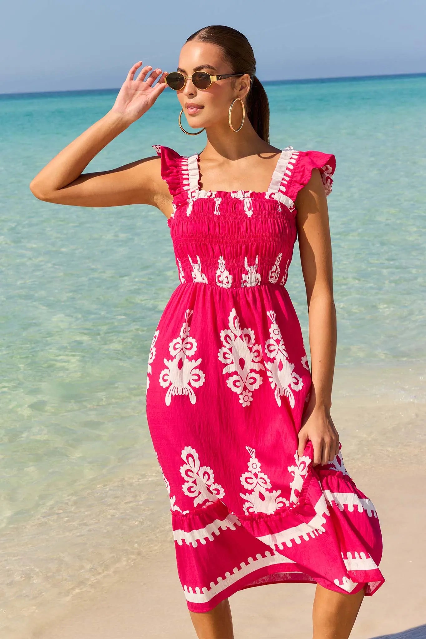 Woman in a pink dress with white patterns standing on a beach.
