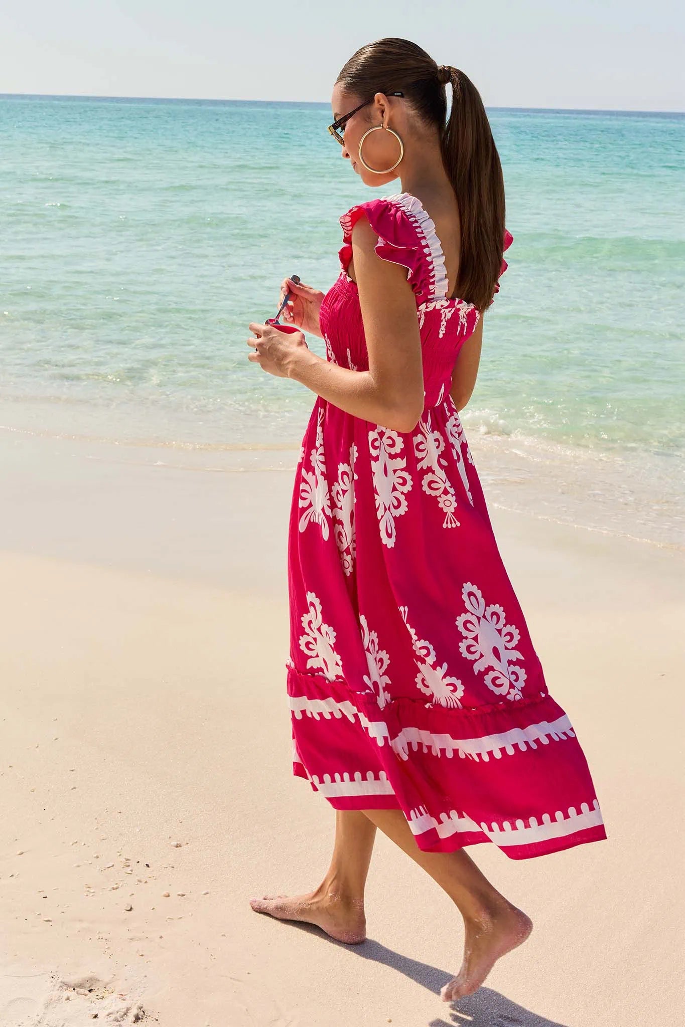 Woman in a red dress with white patterns walking on a beach.