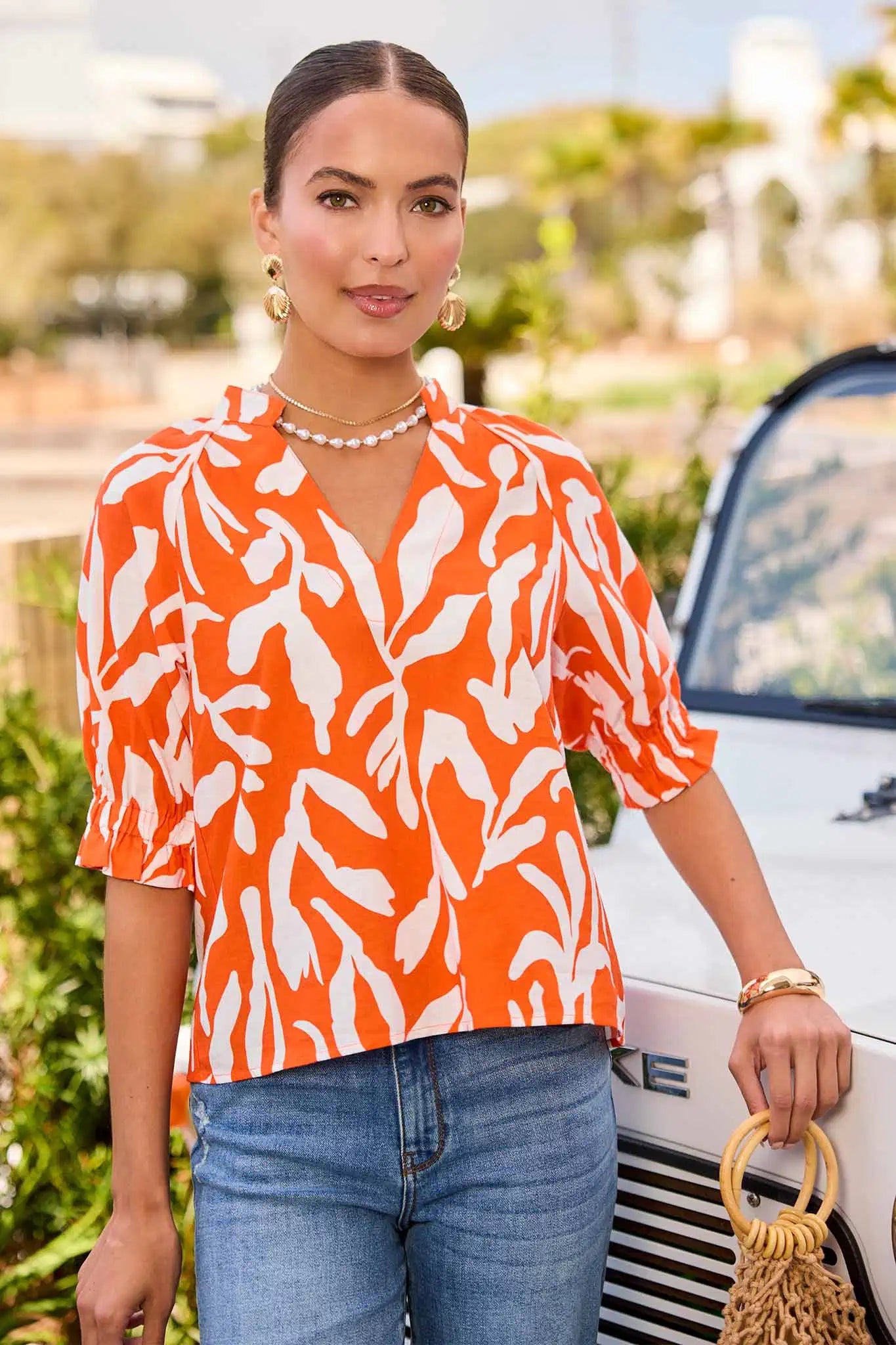 Woman wearing an orange and white patterned shirt with blue jeans, standing next to a white car.
