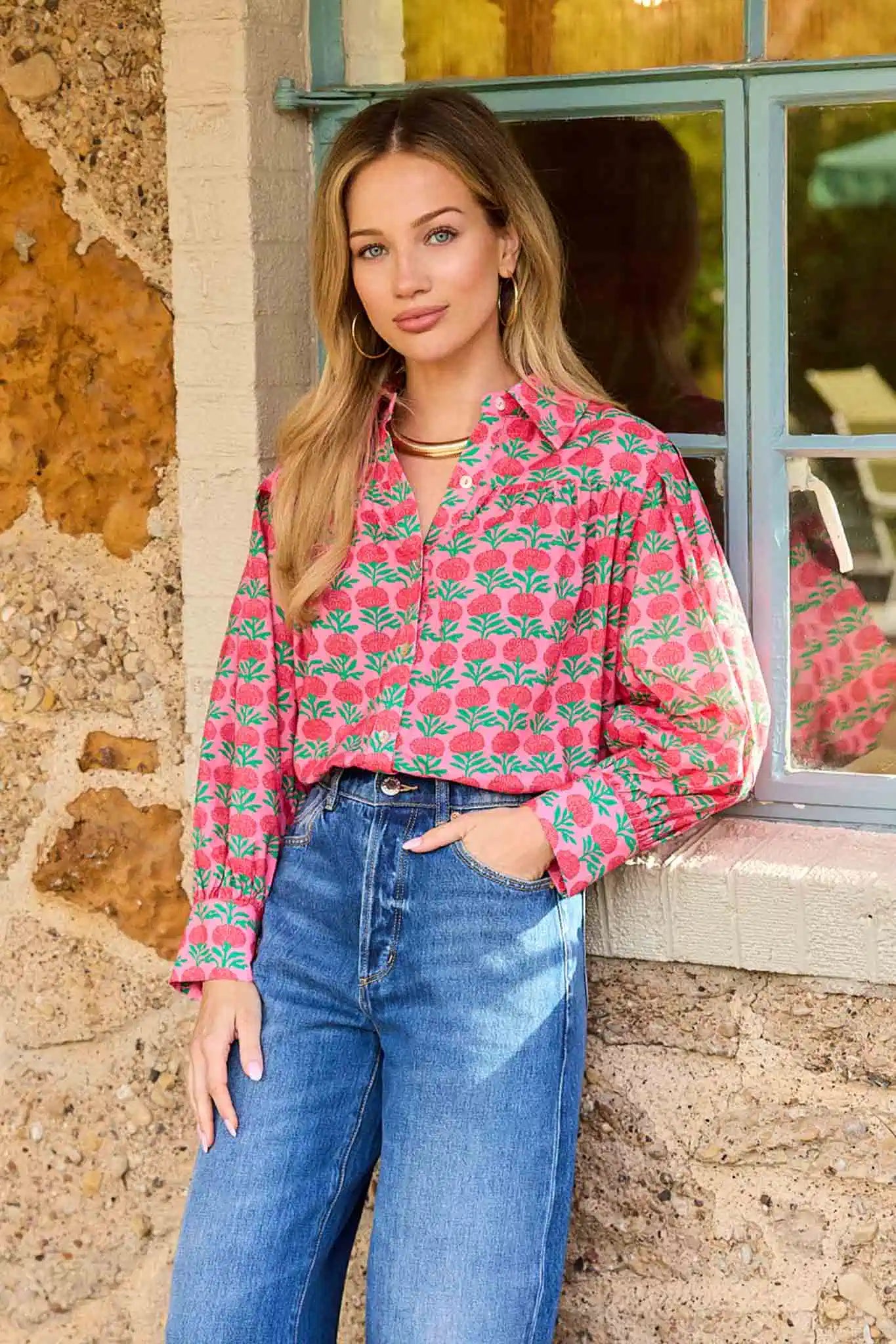 Woman wearing a pink floral blouse and blue jeans standing against a stone wall.