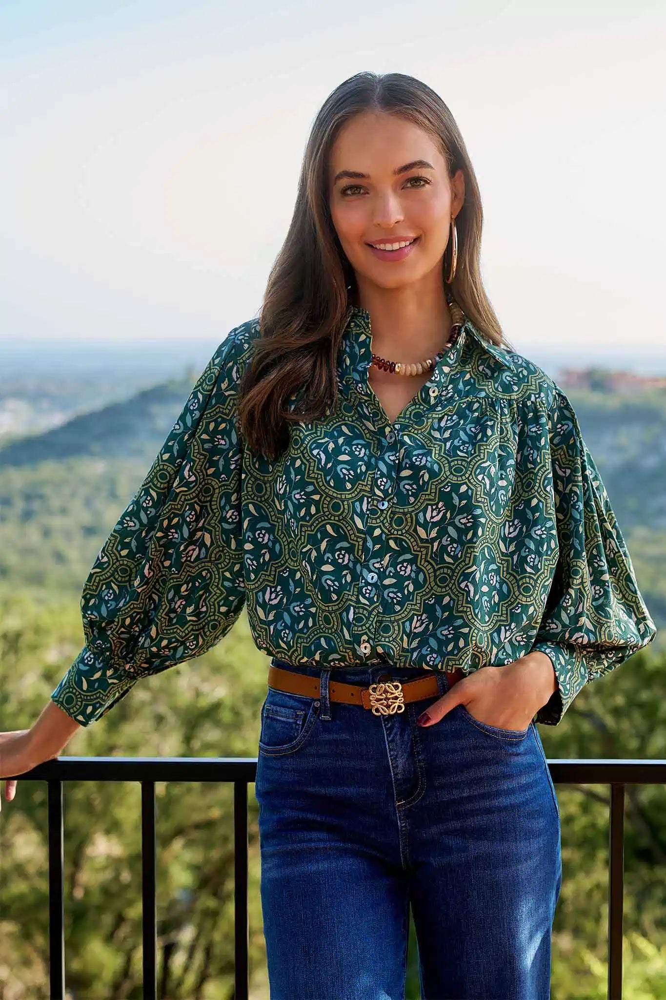 Young woman wearing Trudy Olive Floral Top with ornate print, puffed sleeves, and button-front, paired with blue jeans, enjoying a serene outdoor view.