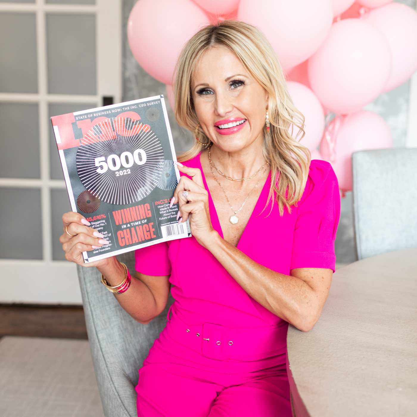 women in pink jumpsuit holding magazine with pink balloons in background