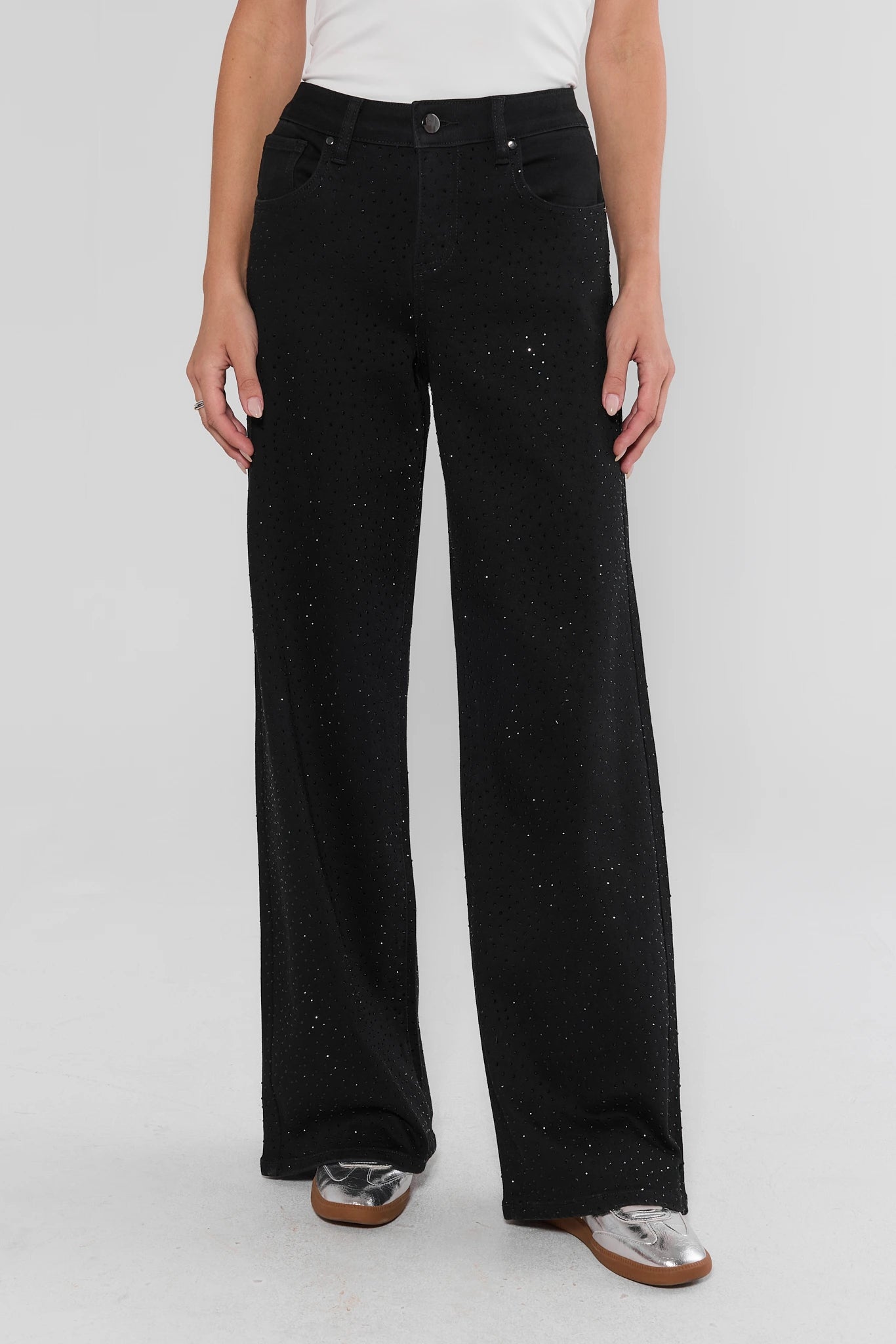 A model wearing a pair of black wide-leg jeans with subtle silver glitter or speckled embellishments, paired with a white top and metallic silver shoes, against a light gray studio background.