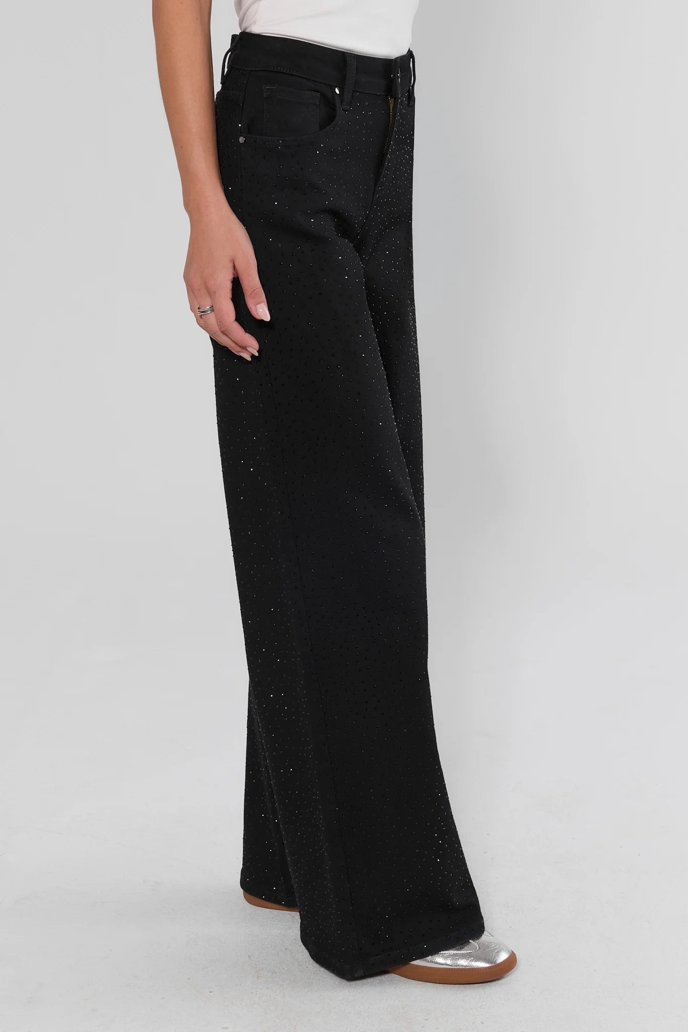 Black wide-leg jeans with subtle silver glitter speckles, shown on a model from the waist down wearing silver flat shoes against a light gray studio background.