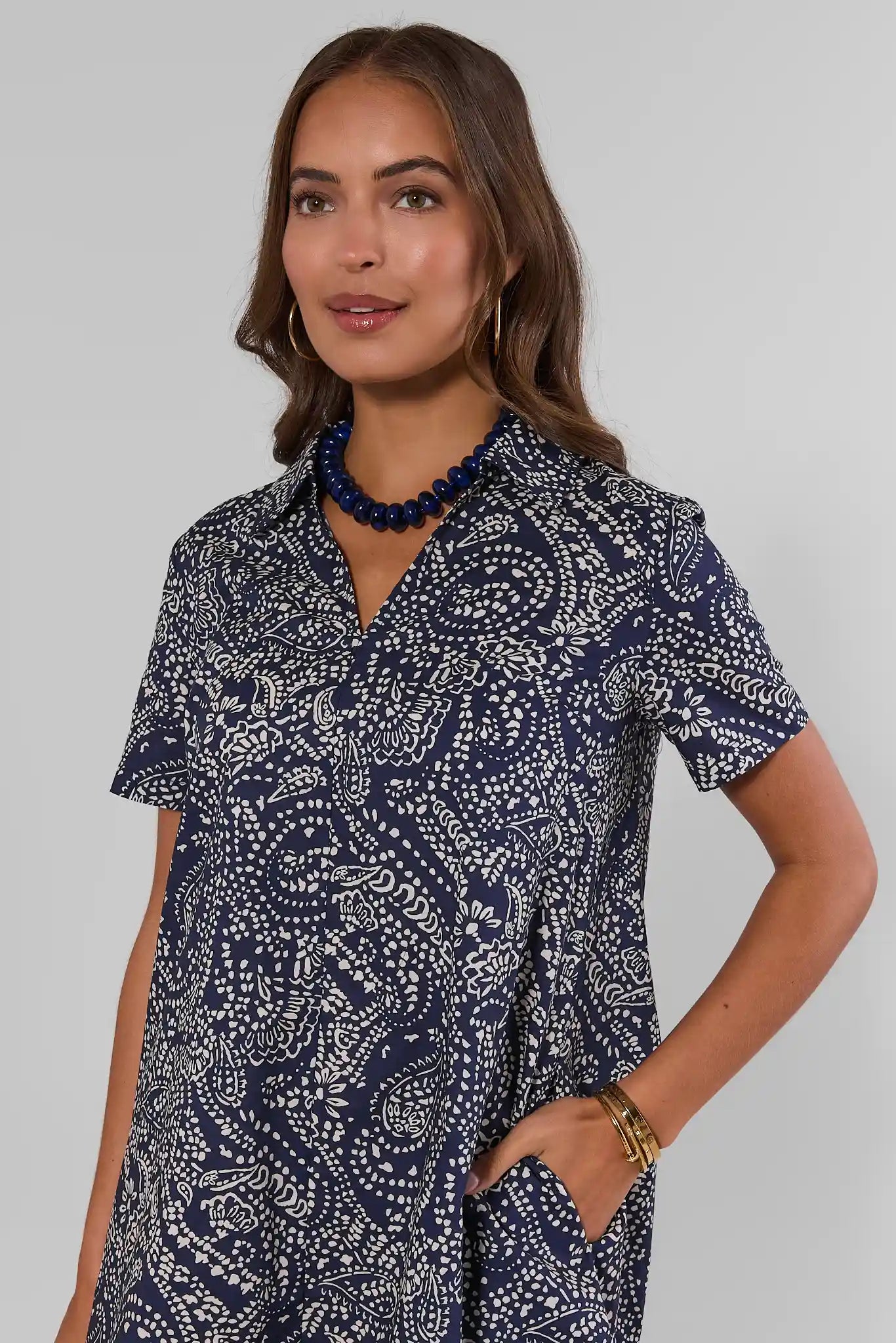 Close up a woman modeling a short-sleeve mini dress with a navy and white paisley pattern, paired with white sneakers and a dark blue beaded necklace, against a light gray studio background.