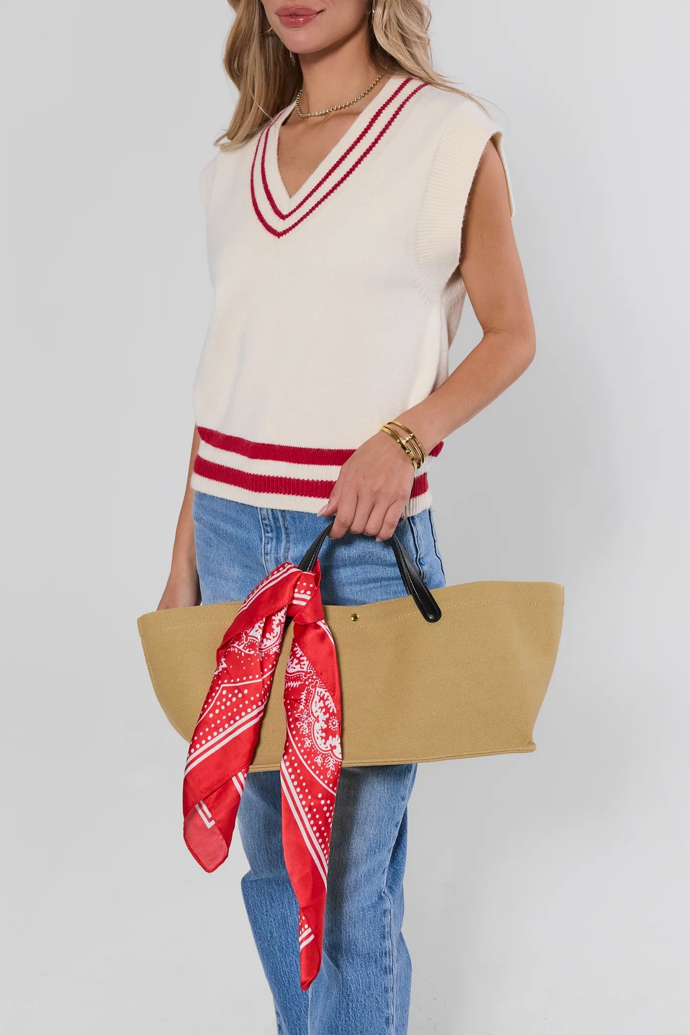 A woman models a cream knit sweater vest with red and white striped trim, paired with blue jeans and holding a tan tote bag with a red patterned scarf.