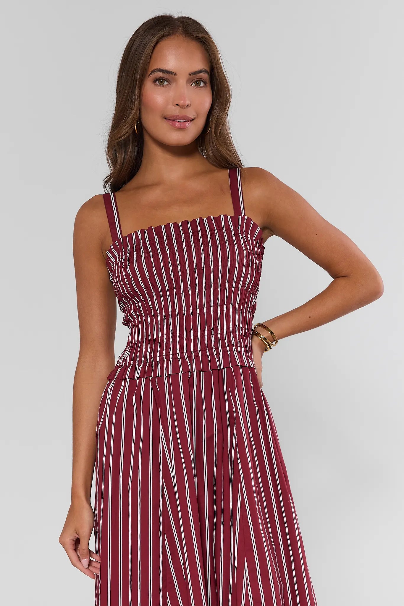 A woman modeling a sleeveless midi dress with thin shoulder straps, a smocked bodice, and vertical red and white stripes, set against a plain light gray studio background.