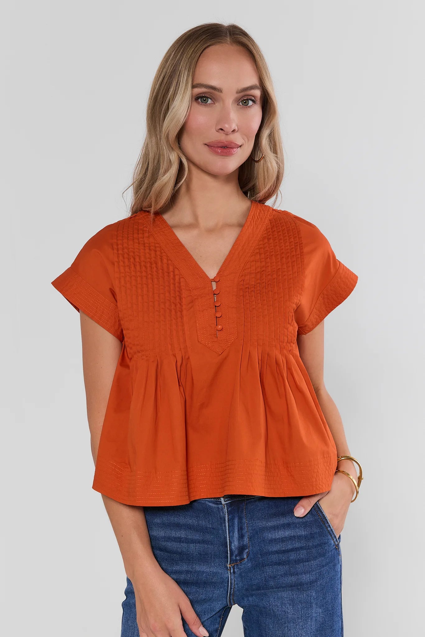 A woman wearing a bright orange short-sleeve peplum top with a V-neck and button placket, paired with blue jeans, against a light gray studio background.