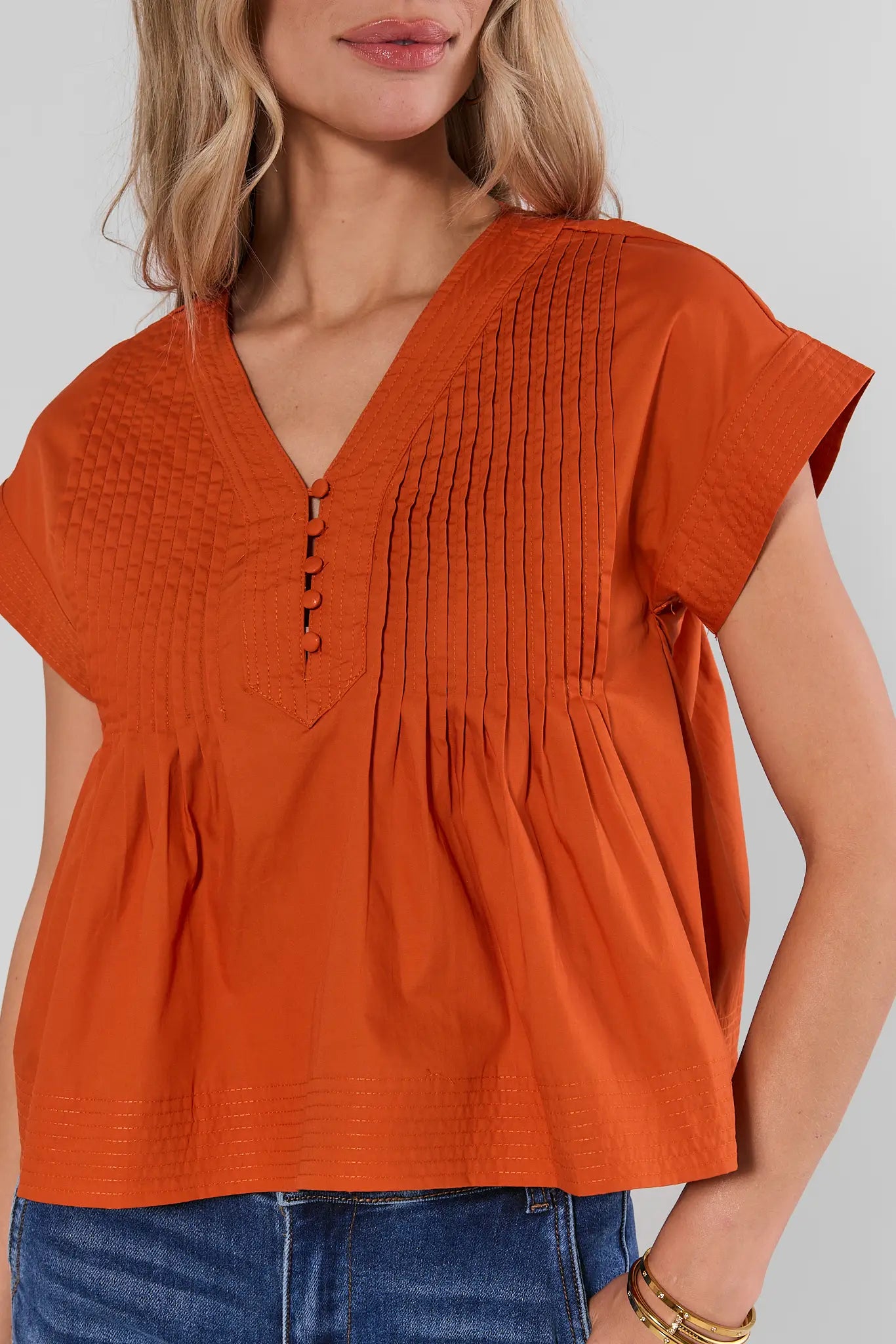 Close up a woman wearing a bright orange short-sleeve peplum top with a V-neck and button placket, paired with blue jeans, against a light studio background.