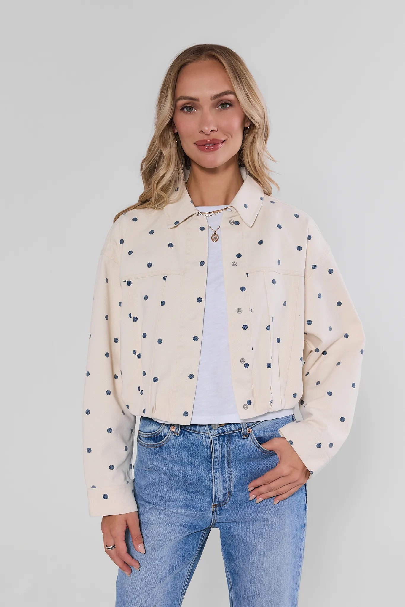 A woman modeling a cropped cream-colored denim jacket with black polka dots, worn over a white t-shirt and blue jeans, against a light gray studio background.