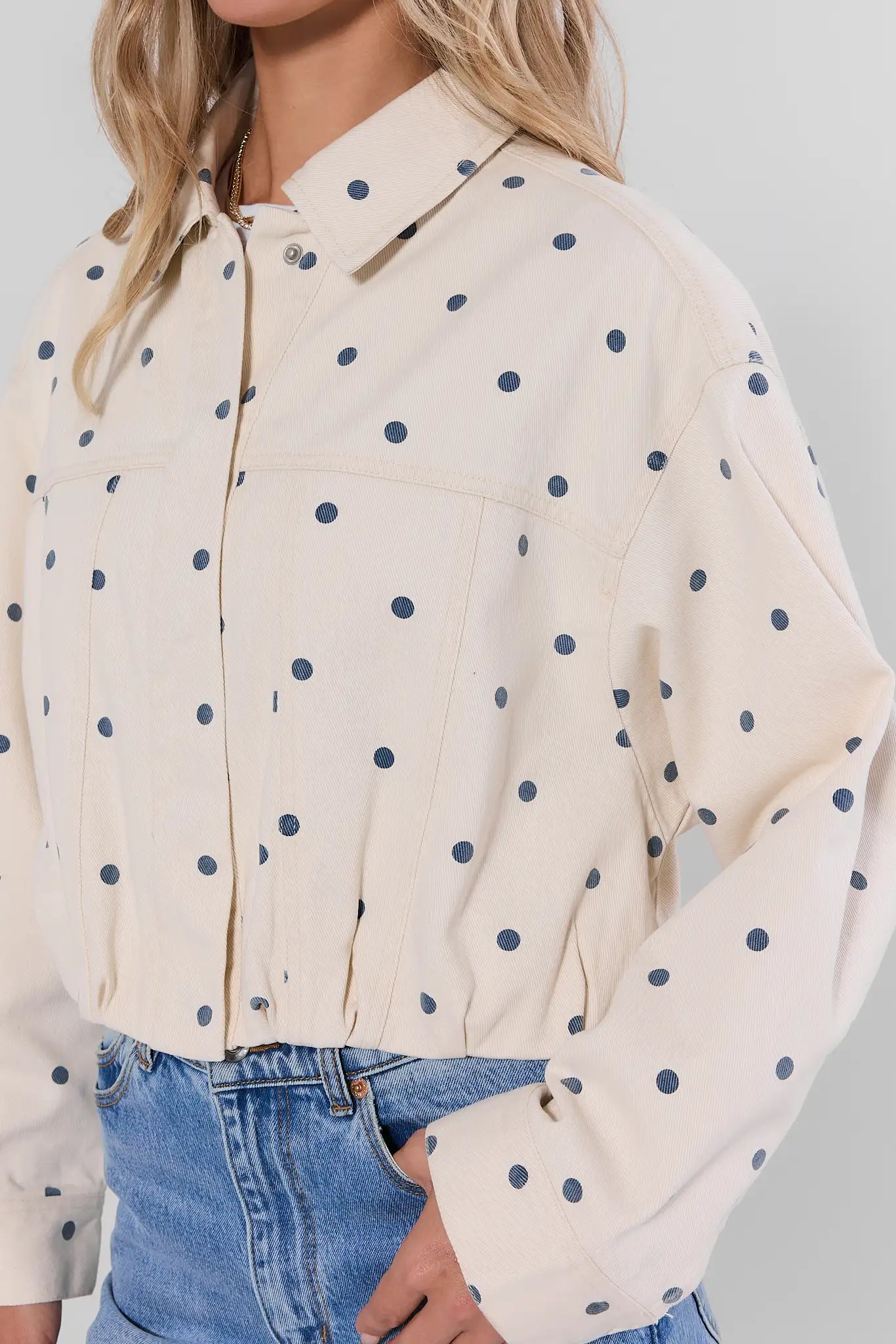 close up A woman wearing a cream-colored long-sleeve button-up shirt with navy blue polka dots, paired with blue jeans.
