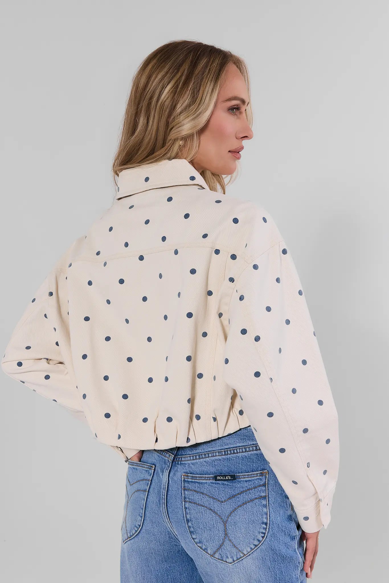 A woman shown from the back wearing a cream-colored long-sleeve button-up shirt with dark navy blue polka dots, paired with blue jeans, against a plain gray background.