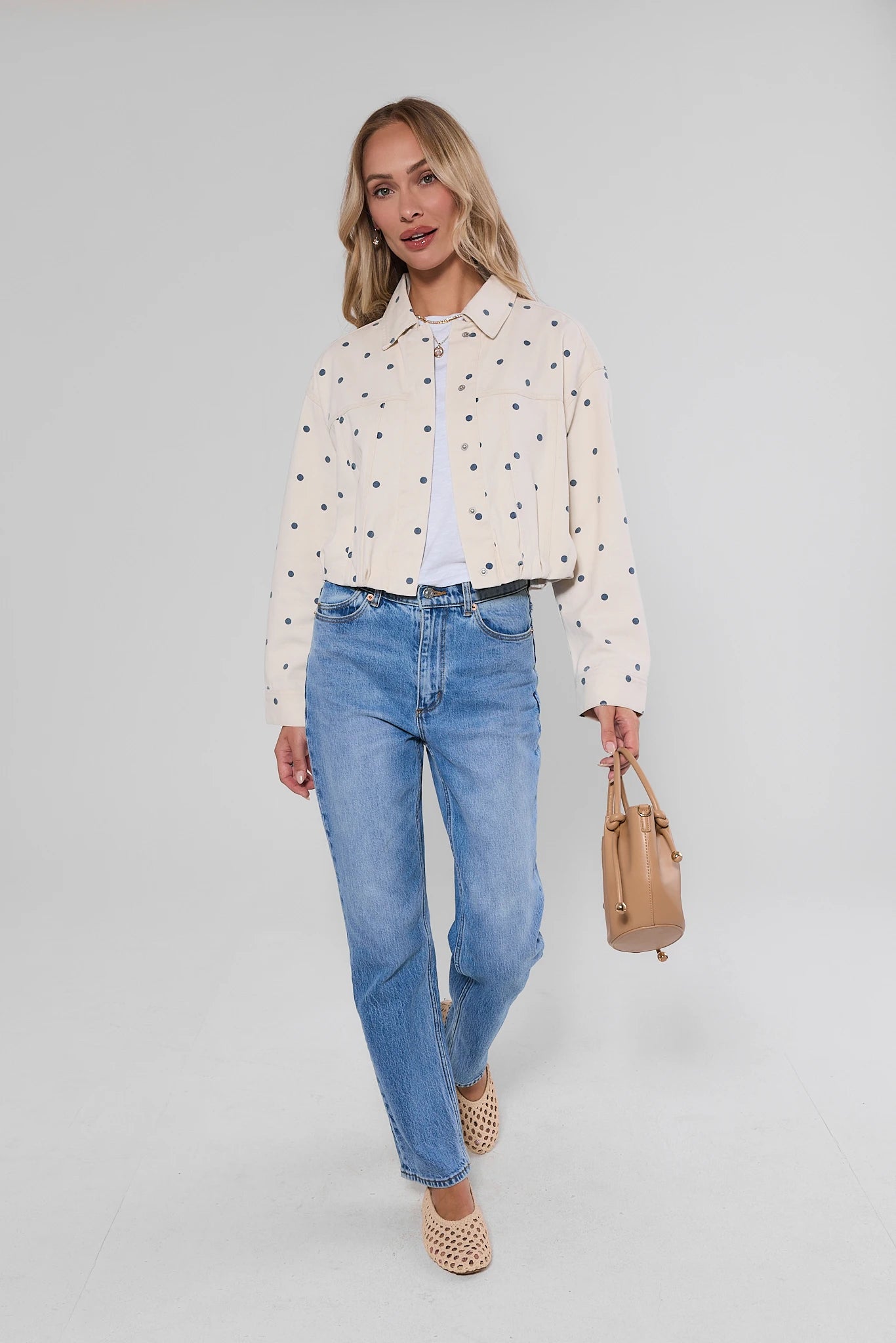A woman modeling a cream cropped jacket with small black embroidered motifs, paired with light blue jeans, a white t-shirt, and holding a tan handbag against a light gray studio background.