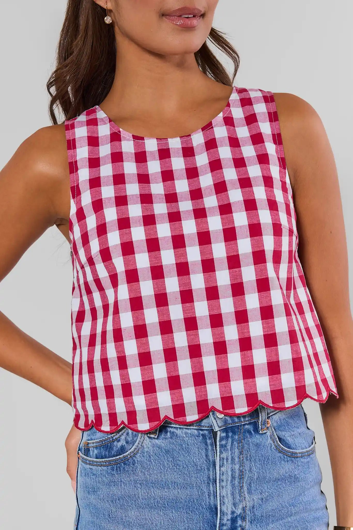 Close up a woman wearing a sleeveless red and white gingham check top with a scalloped hem, paired with blue jeans, against a light gray background.