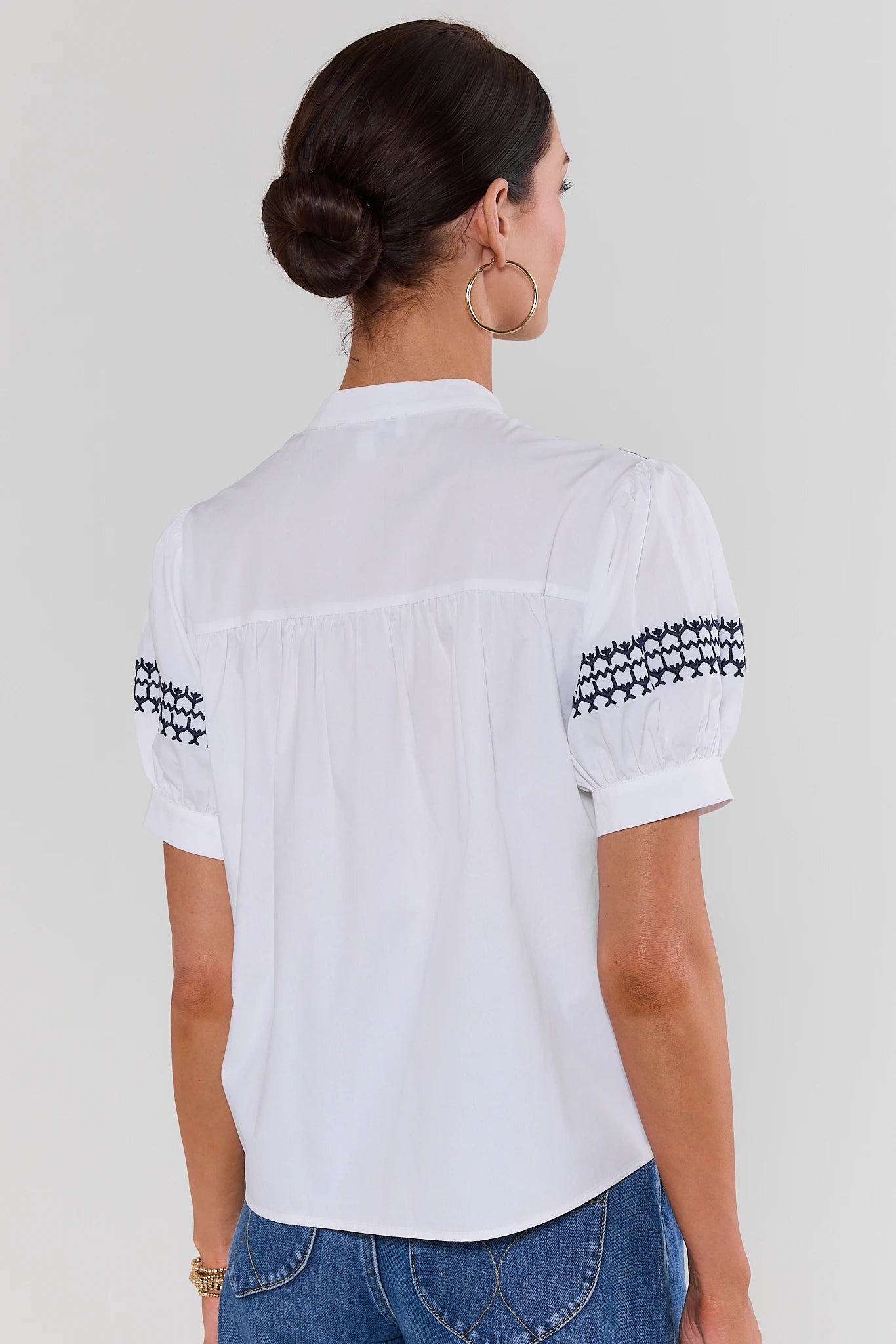 Woman standing wearing a white blouse with black patterns on sleeves against a plain background