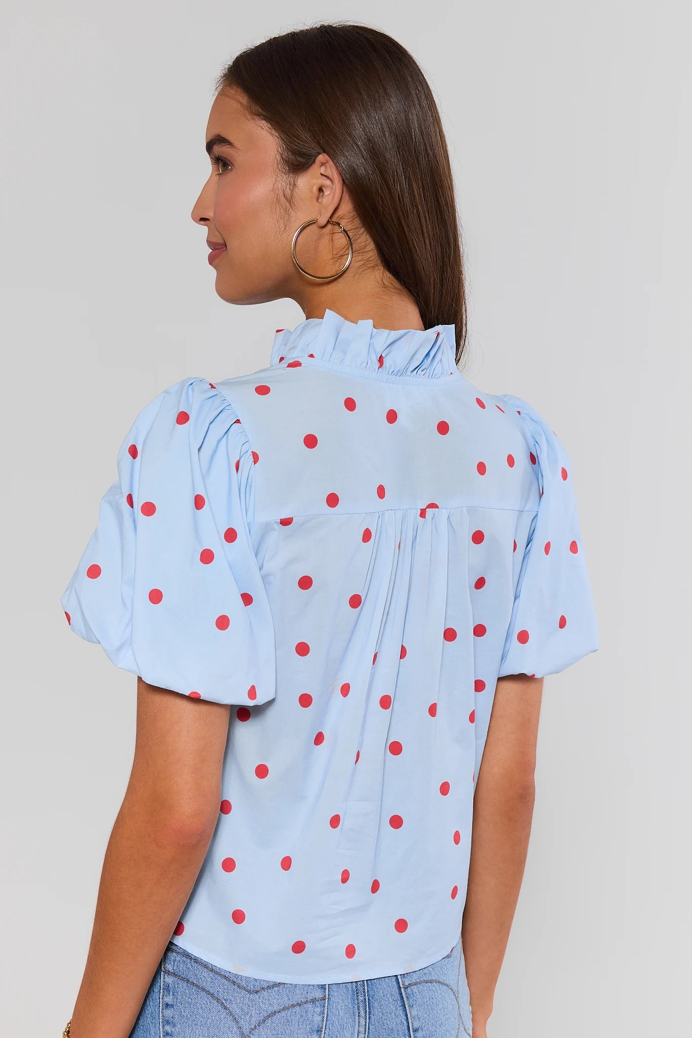 Woman standing wearing a light blue blouse with red polka dots on a plain background