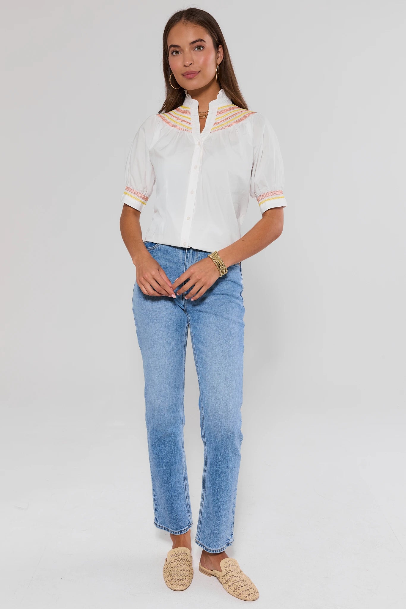 Woman standing wearing a white blouse with yellow accents and blue jeans on a plain background