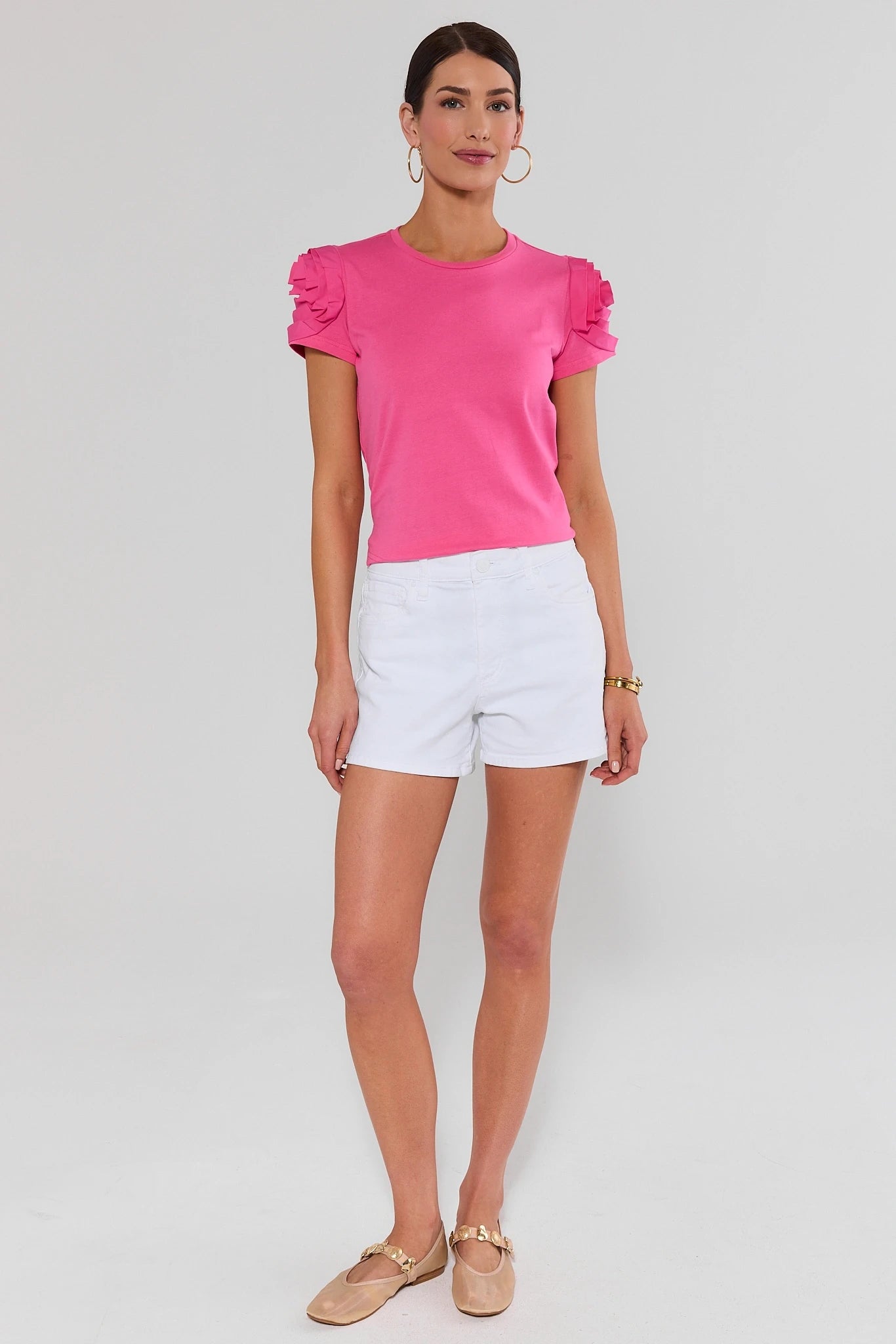 Woman wearing a bright pink top with ruffled sleeves and white shorts on a light gray background