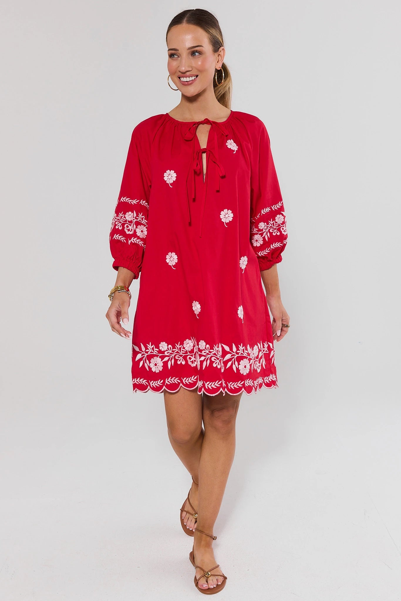 Woman standing wearing a red dress with white floral embroidery on a white background