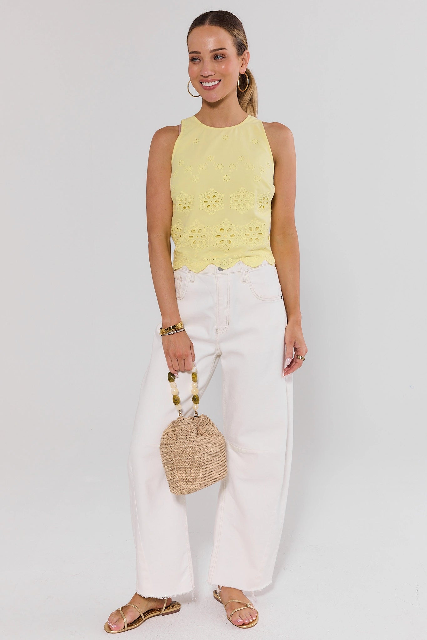 Woman wearing a yellow sleeveless top and white pants, holding a straw bag on a white background