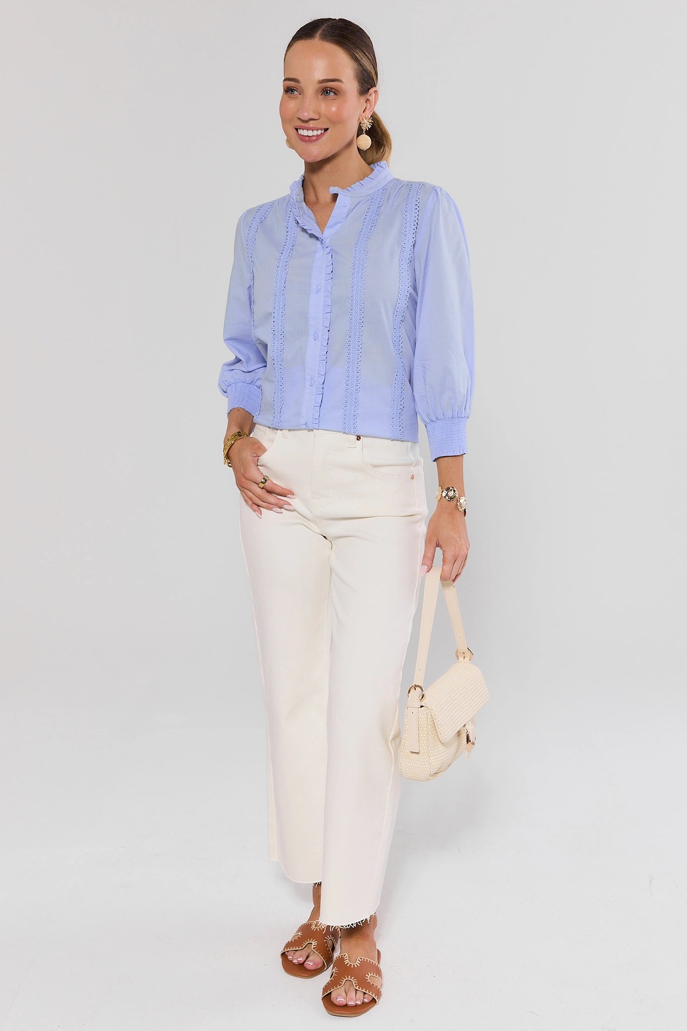 Woman wearing a light blue shirt and white pants with a beige handbag on a plain background