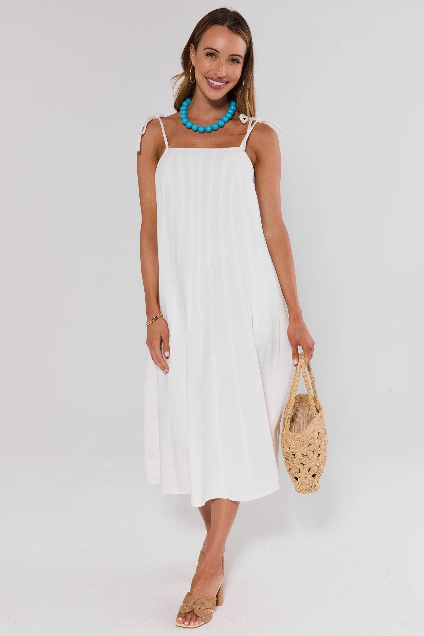Woman wearing a white dress with a blue necklace and holding a woven bag on a white background