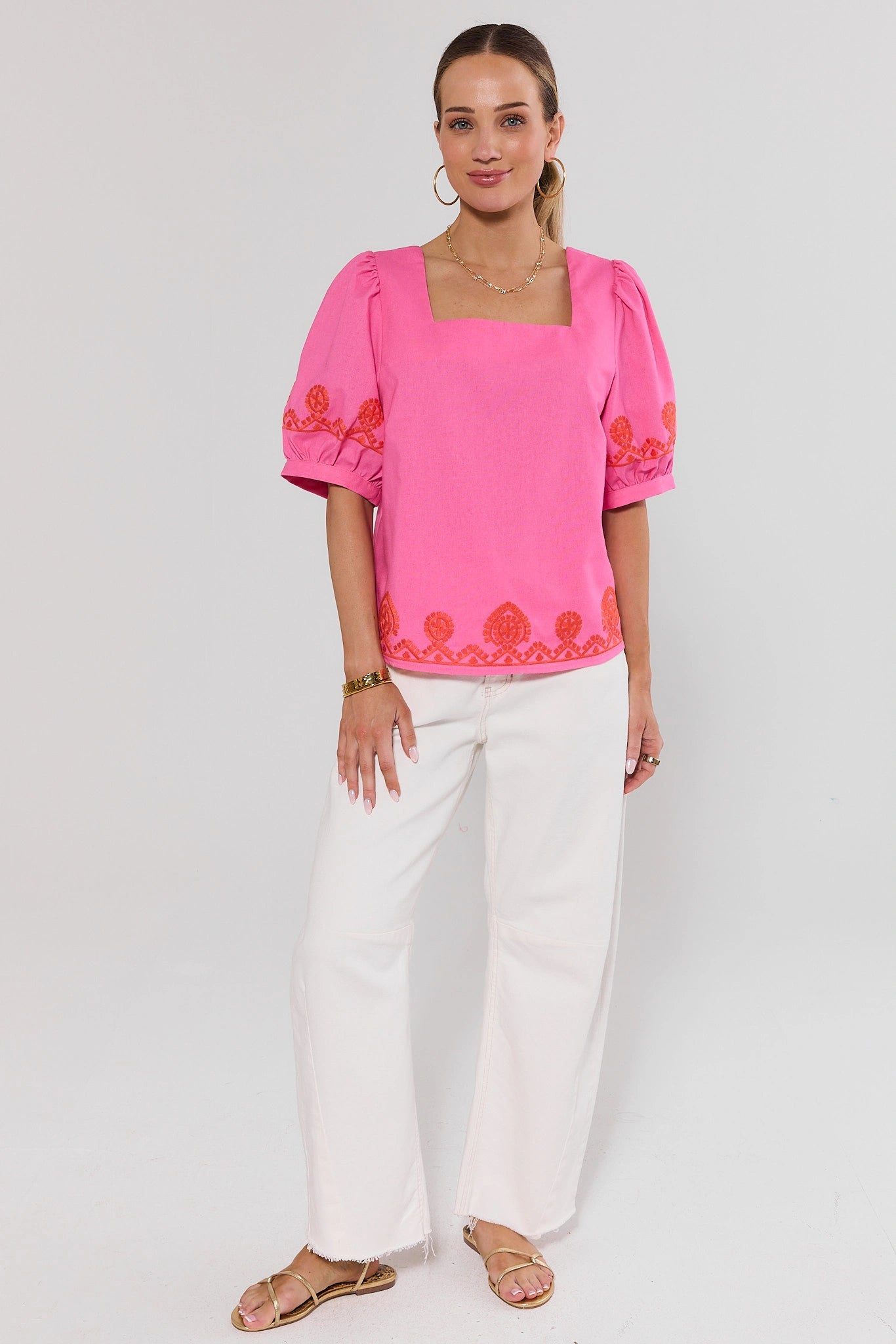 Woman wearing a pink blouse with floral embroidery and white pants on a light gray background