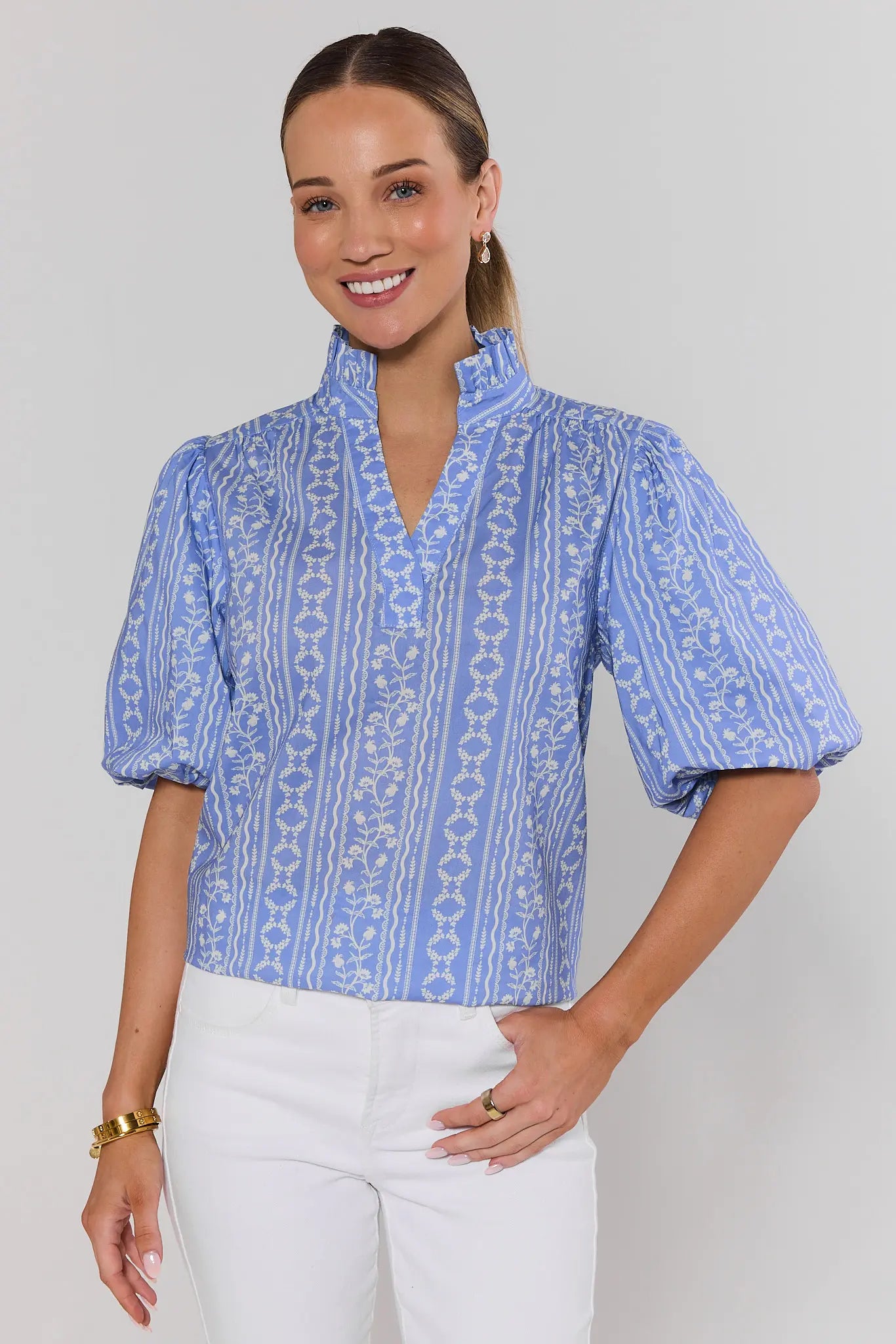 Woman wearing a blue janet patterned blouse with white jeans on a plain background