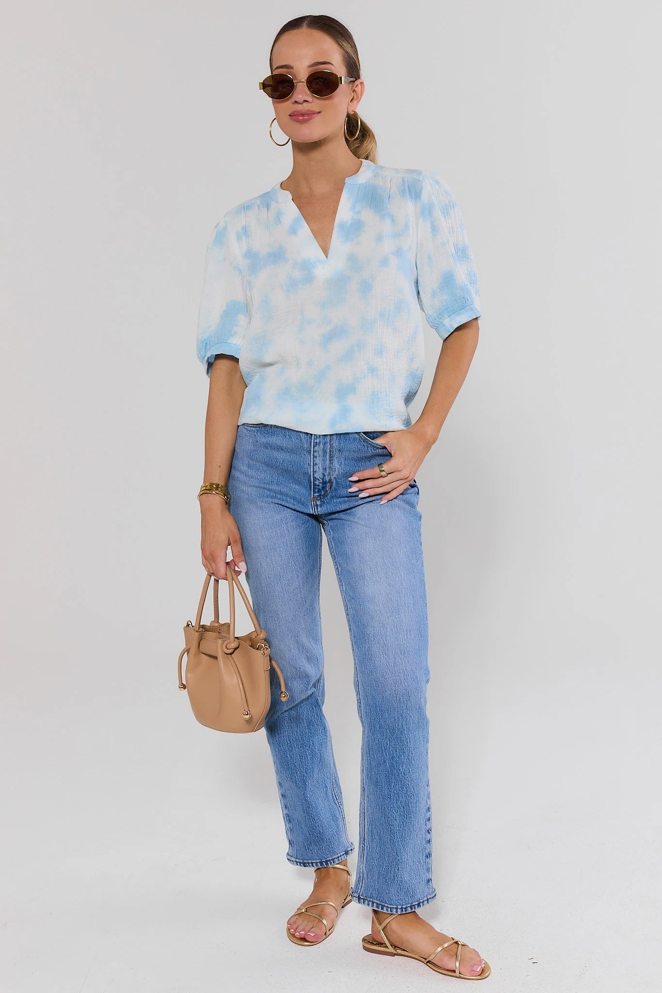 Woman wearing a light blue tie-dye shirt and jeans, holding a beige handbag against a white background