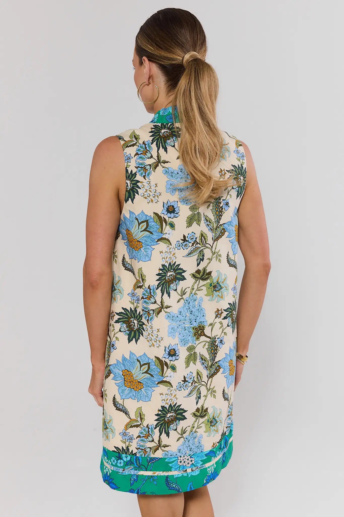 Woman wearing a sleeveless floral dress with blue and green flowers on a plain background standing