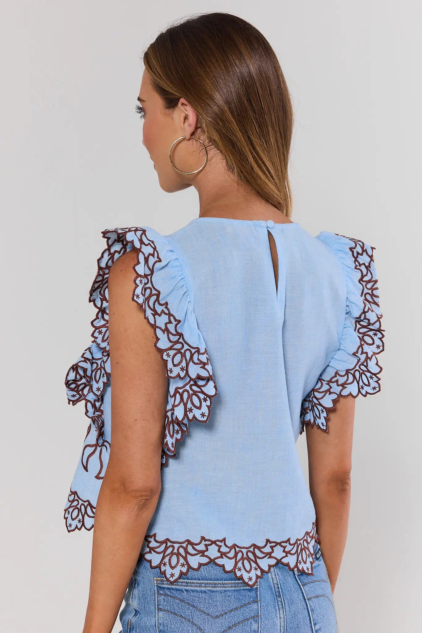 Woman standing wearing a light blue top with floral lace details on a plain background