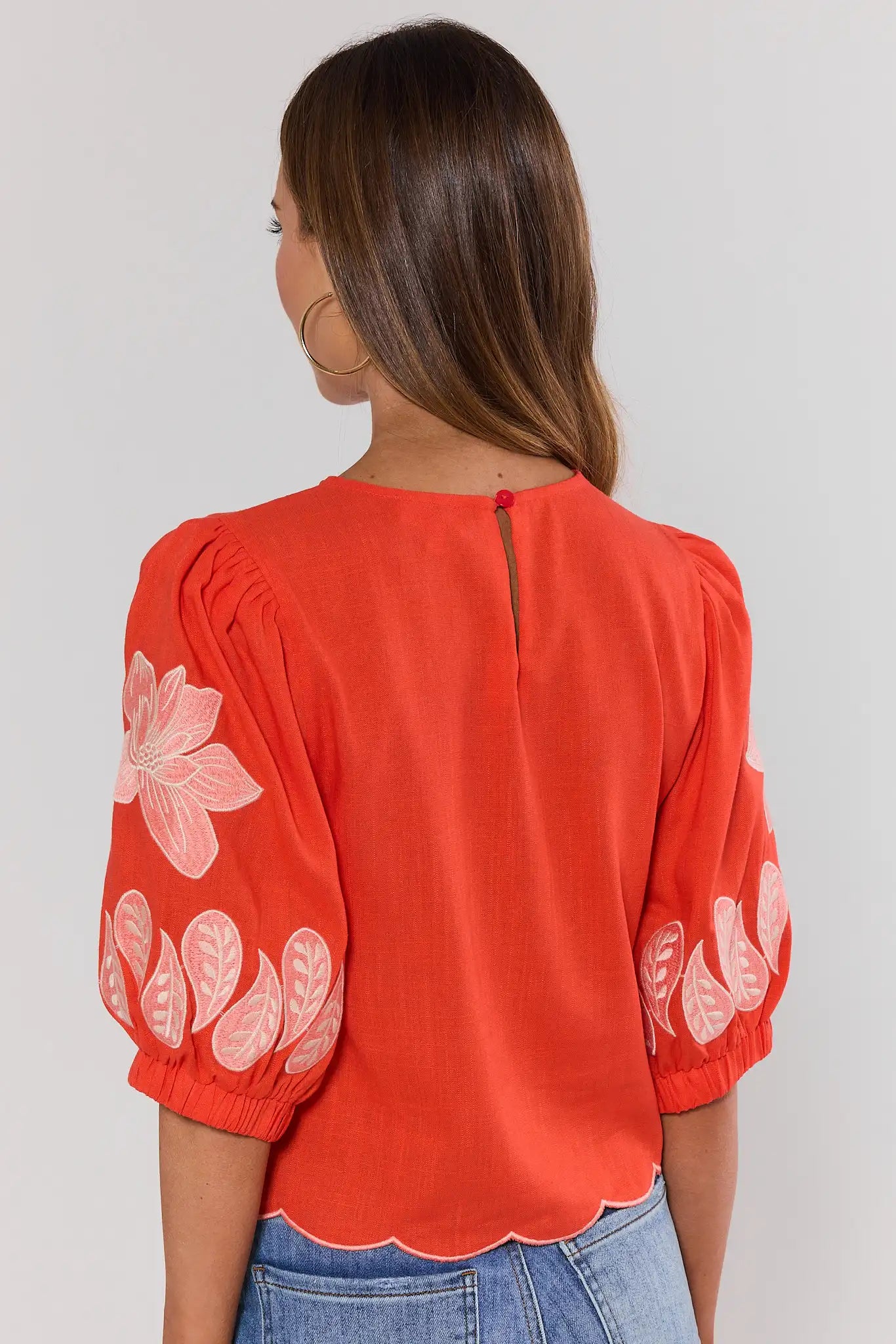 Woman standing wearing a orange blouse with floral patterns on a plain background