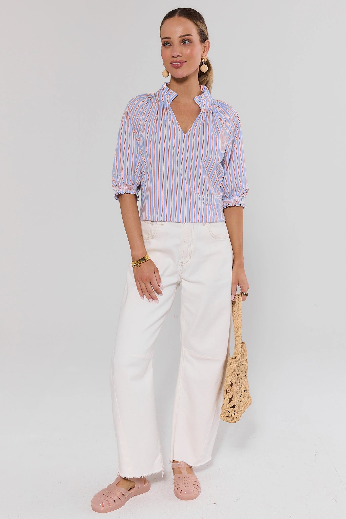 Woman wearing a striped shirt and white pants with a beige bag on a light gray background