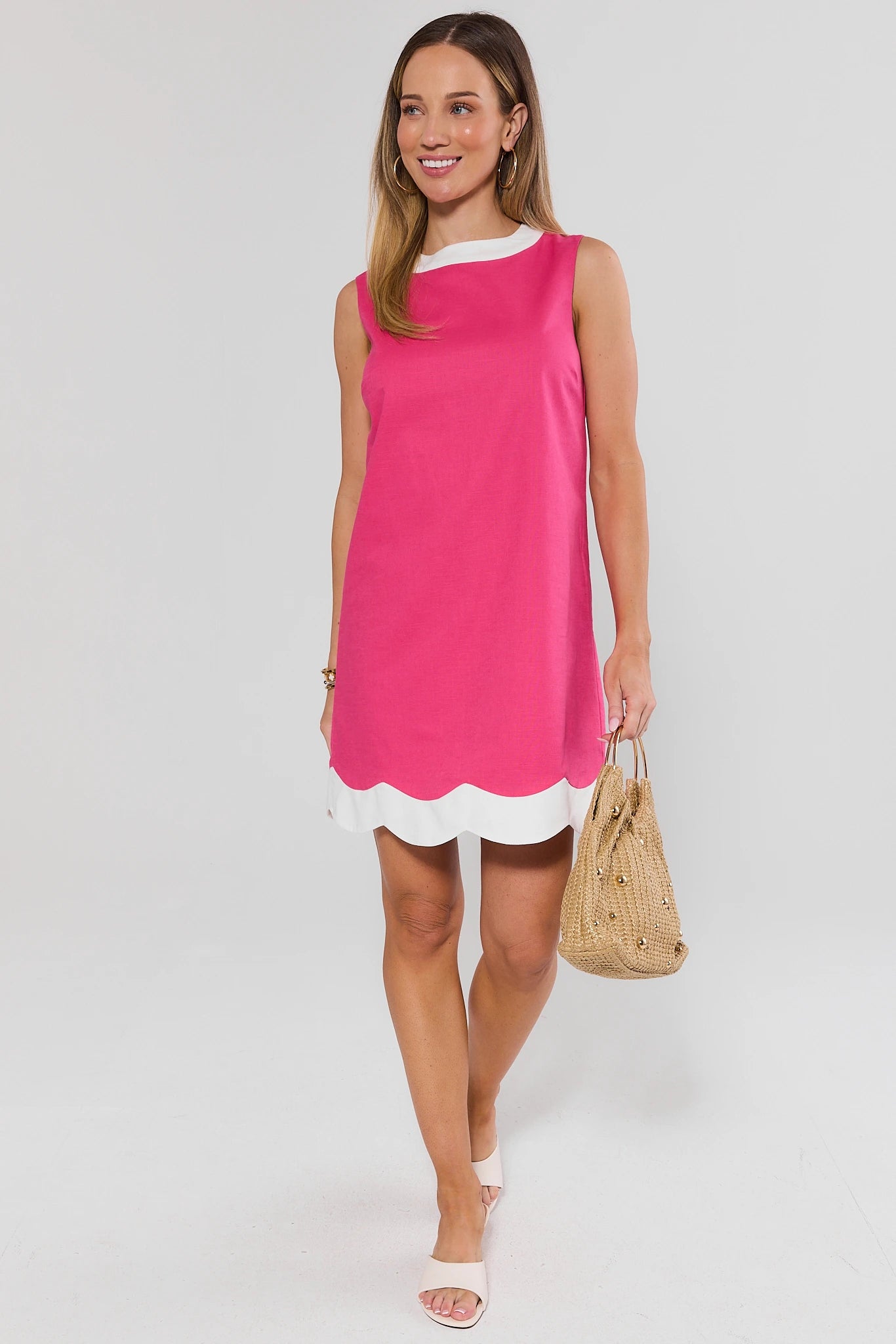 Woman wearing a pink dress with scalloped hem and white shorts, holding a straw bag on a white background