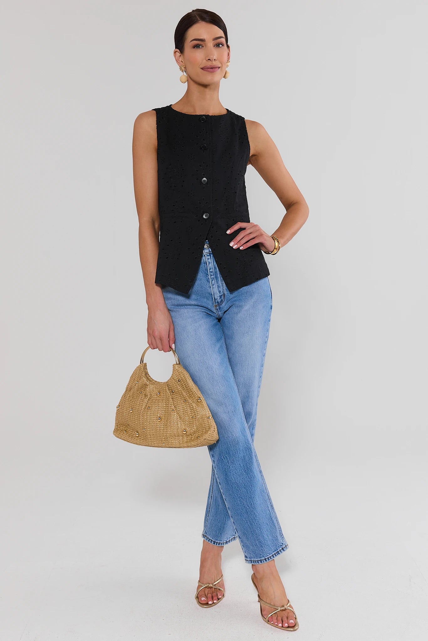Woman wearing a black sleeveless vest and blue jeans, holding a straw bag on a white background