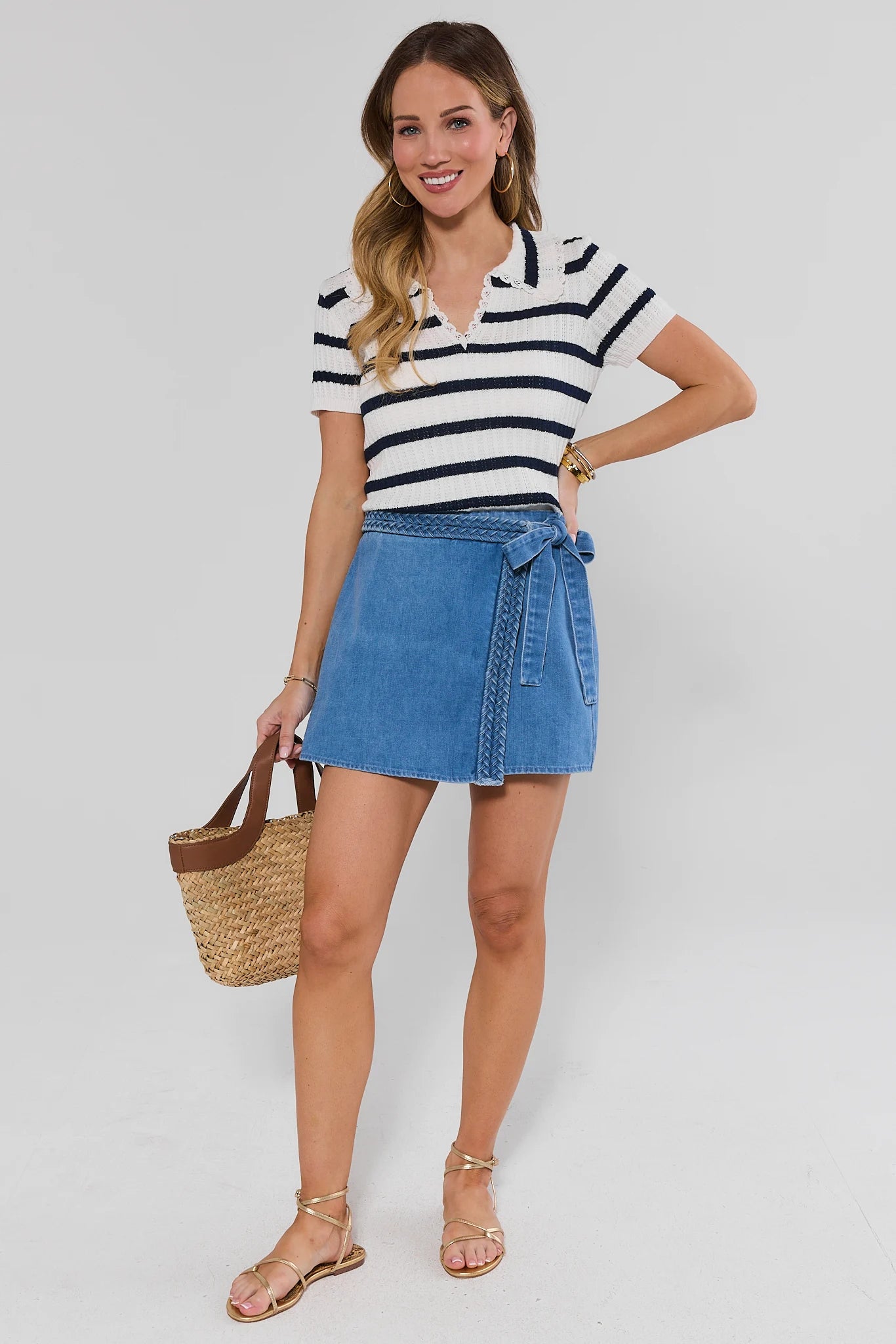 Woman wearing a striped shirt and denim skirt holding a straw bag on a plain background