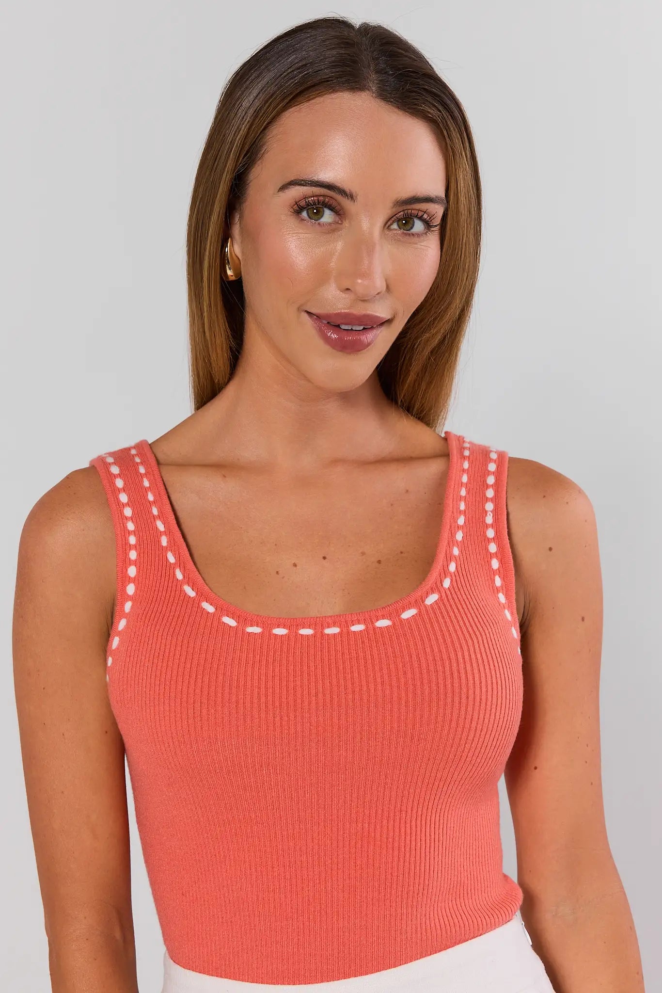 Woman wearing a coral tank top with white stitching on a plain background close up