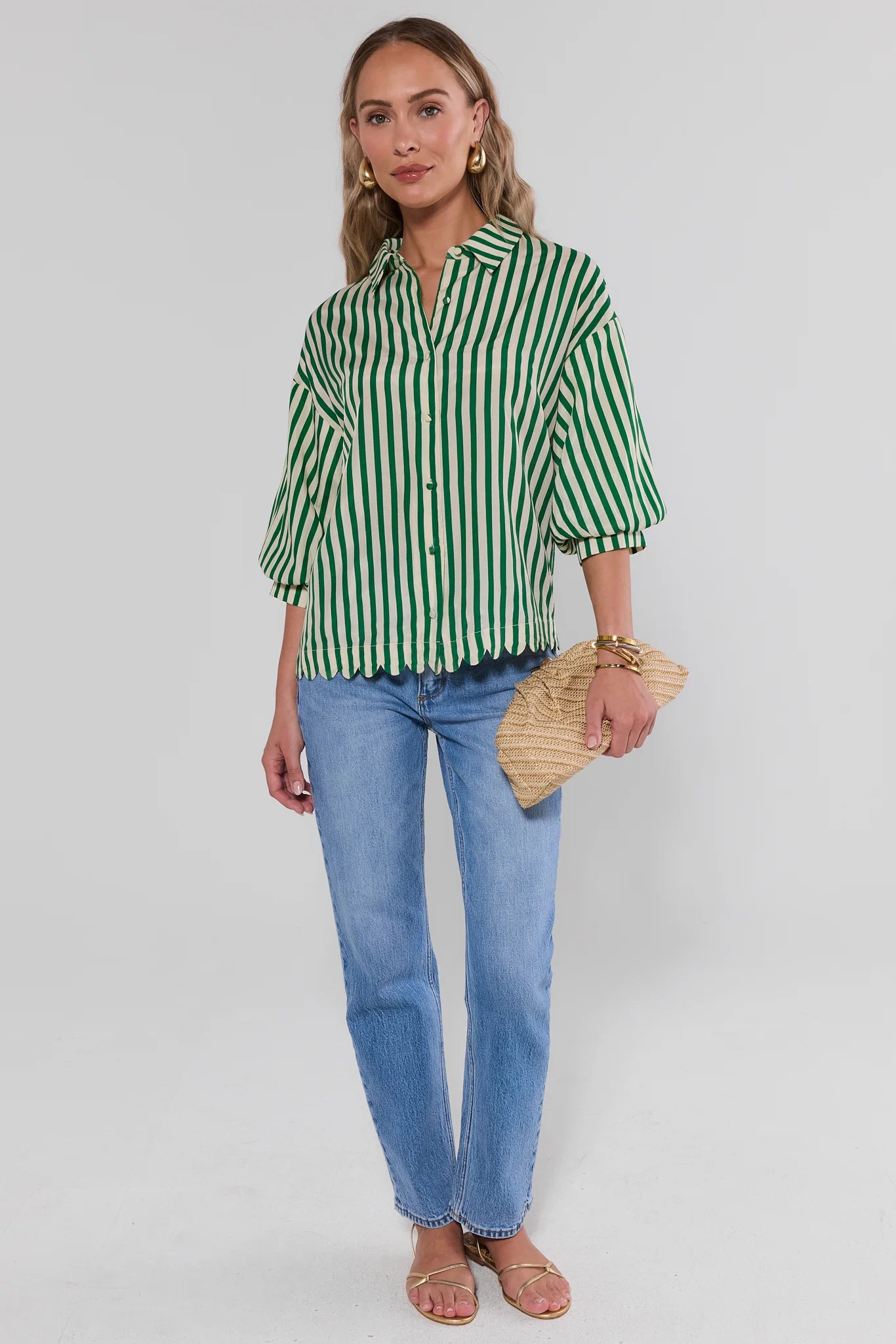 Woman wearing a green and white striped shirt with blue jeans, holding a straw bag on a plain background