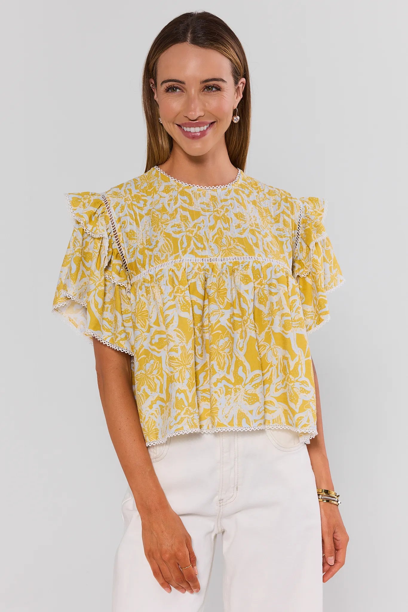 Woman standing wearing a yellow floral blouse with white pants on a light gray background
