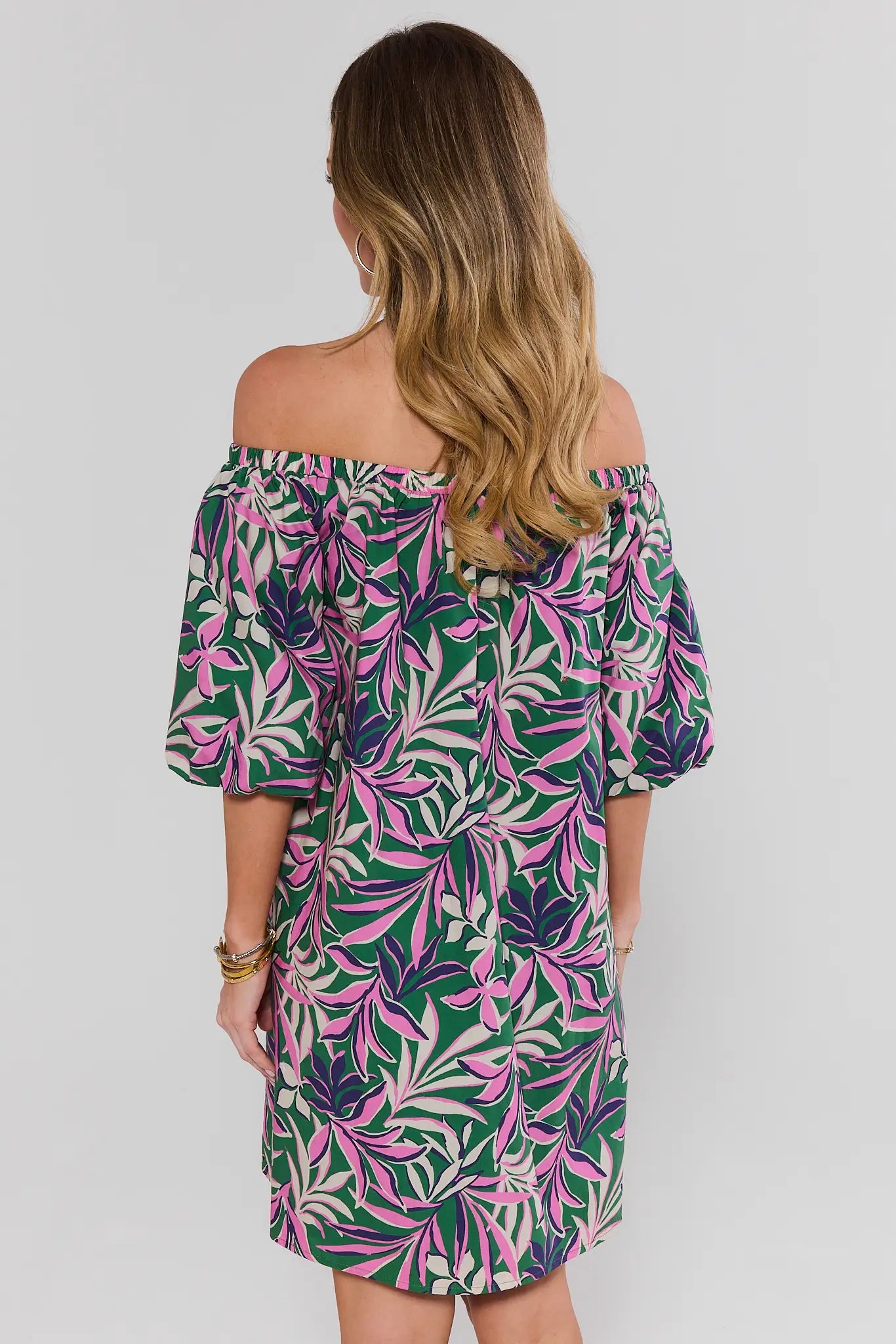 Woman standing wearing a colorful off-shoulder dress with a leaf pattern on a plain background