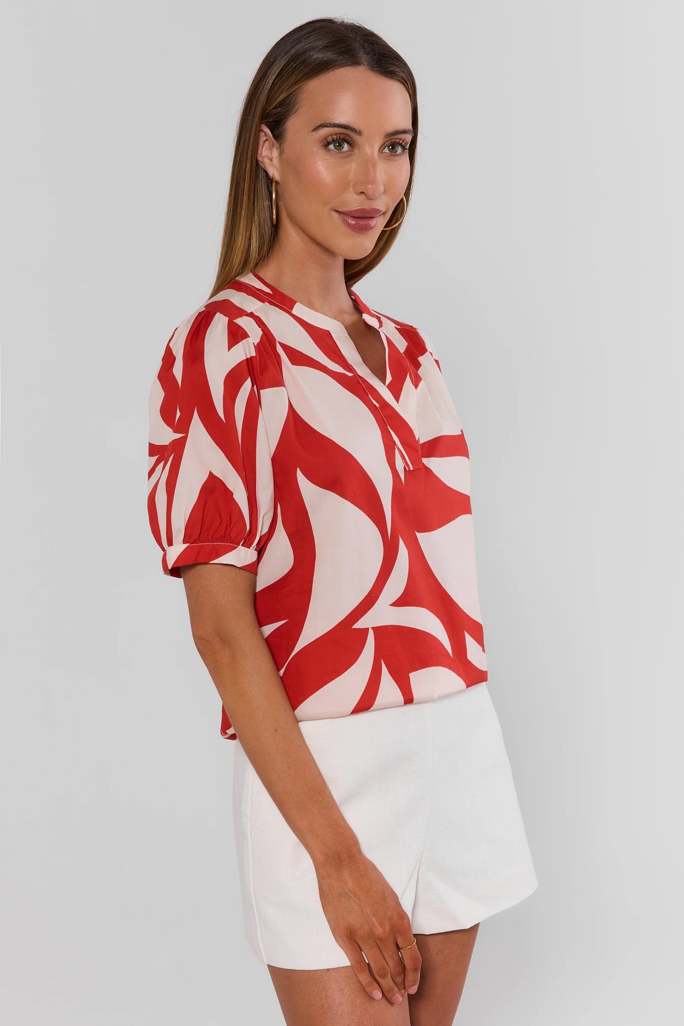 Woman wearing a red and white patterned top with white shorts on a plain background side