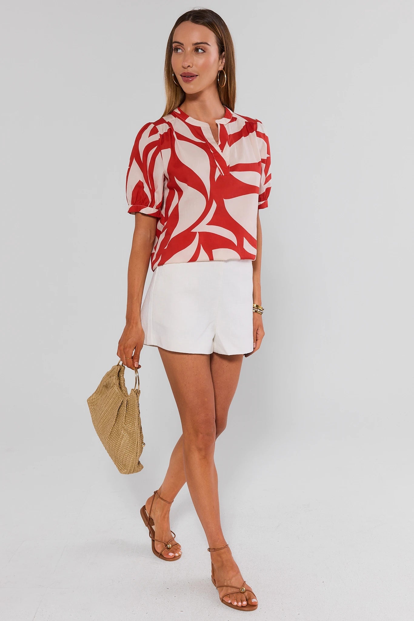 Woman wearing a red and white patterned top with white shorts, holding a beige handbag on a plain background