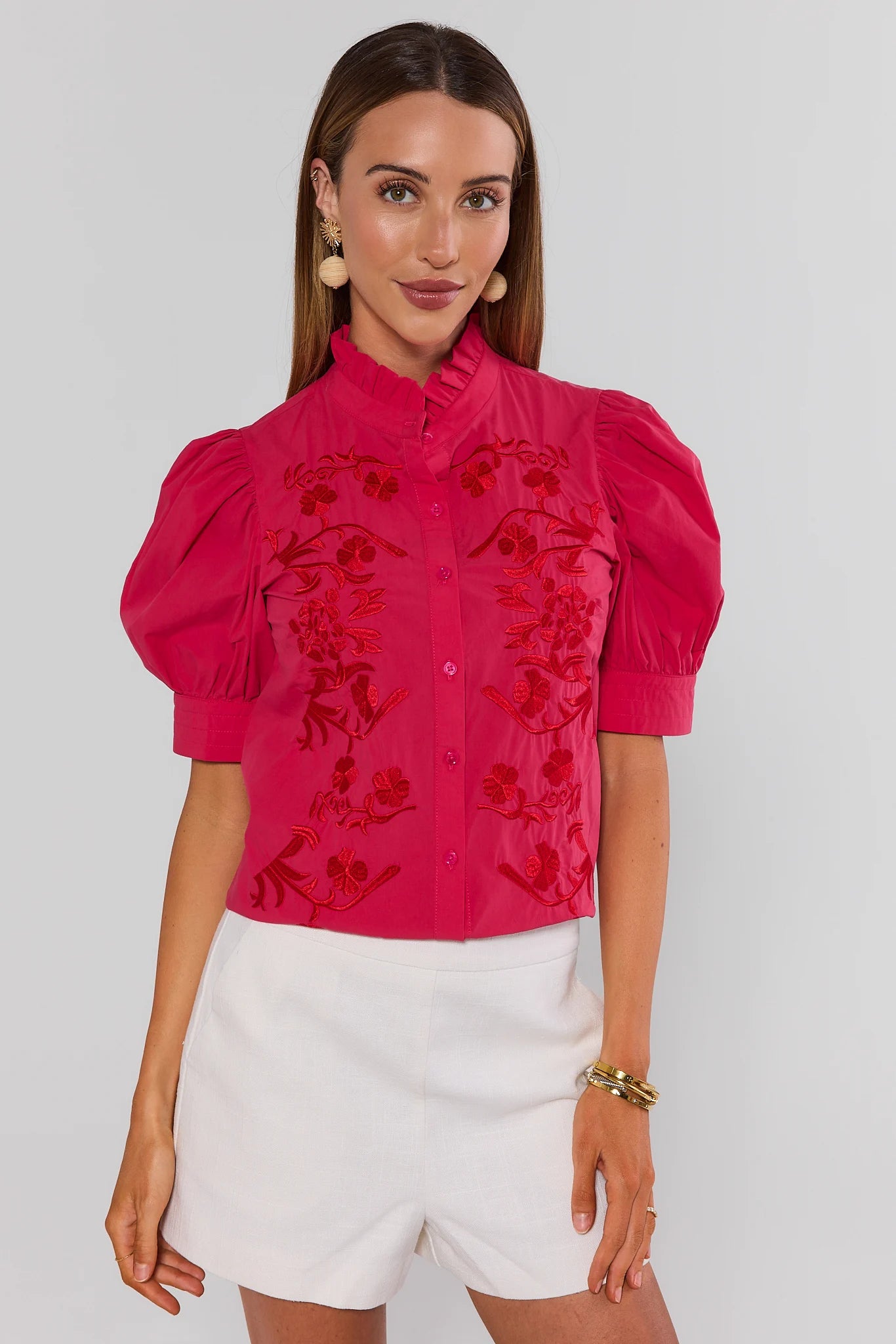 Woman wearing a hot pink embroidered blouse with white shorts on a plain background