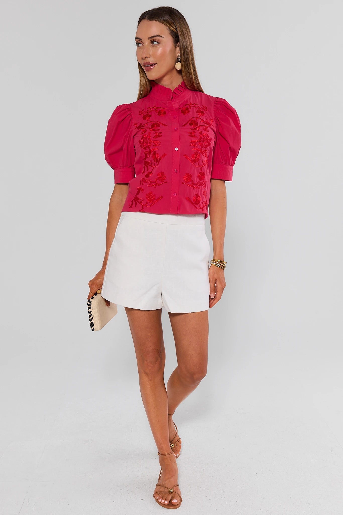 Woman wearing a pink embroidered blouse and white shorts on a light gray background standing