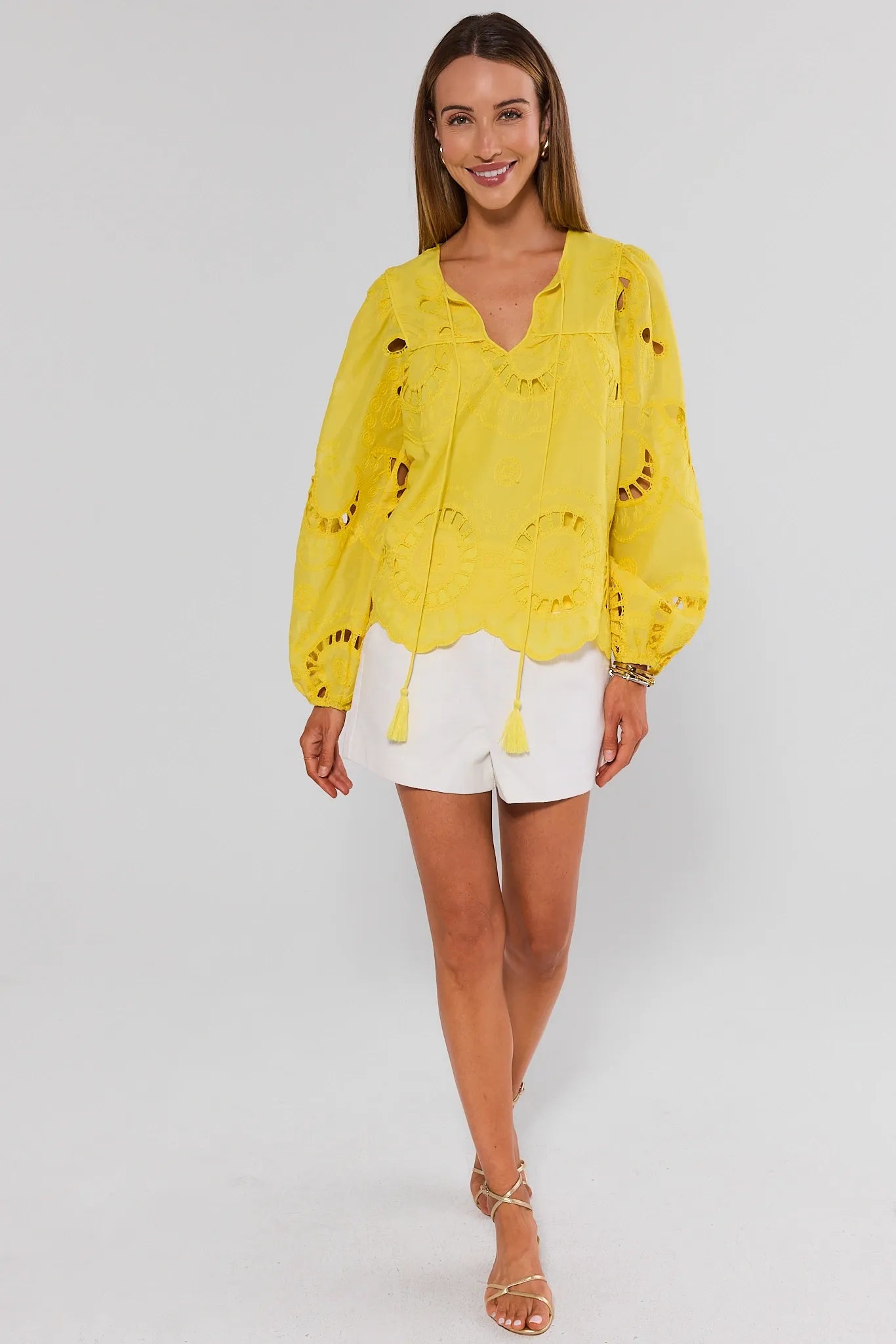 Woman wearing a yellow blouse with floral patterns and white shorts on a plain background standing