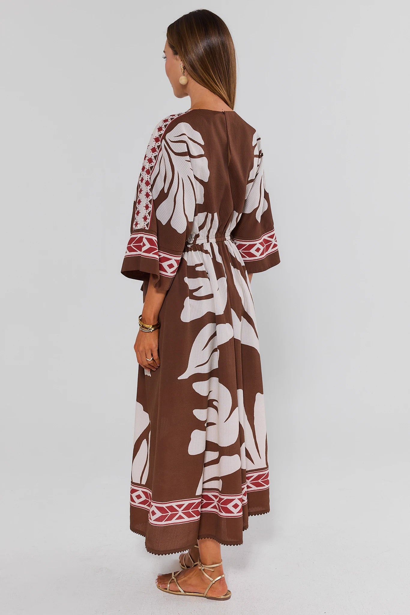 Woman standing wearing a long brown dress with white floral patterns on a plain background